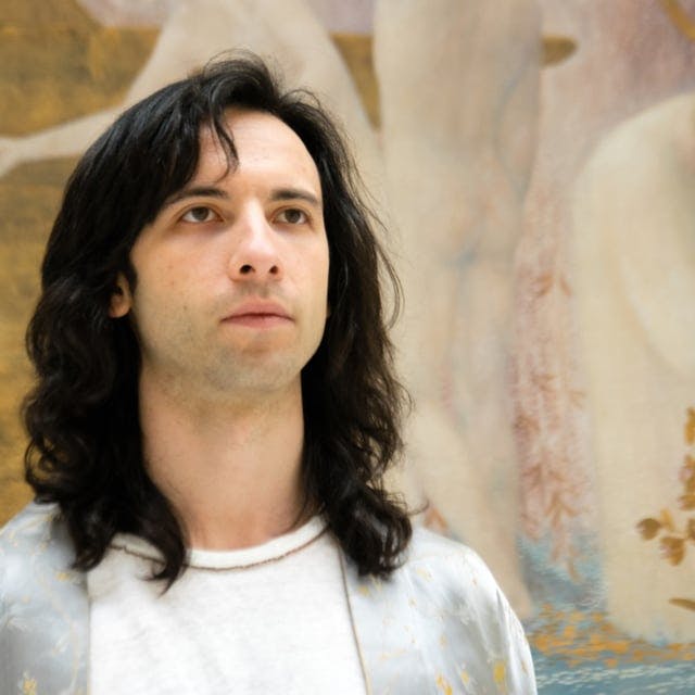 Half-bust of Pietro Marullo facing the camera. He is wearing a light-coloured shirt and is looking slightly upwards. Behind him can be seen part of a Renaissance painting.