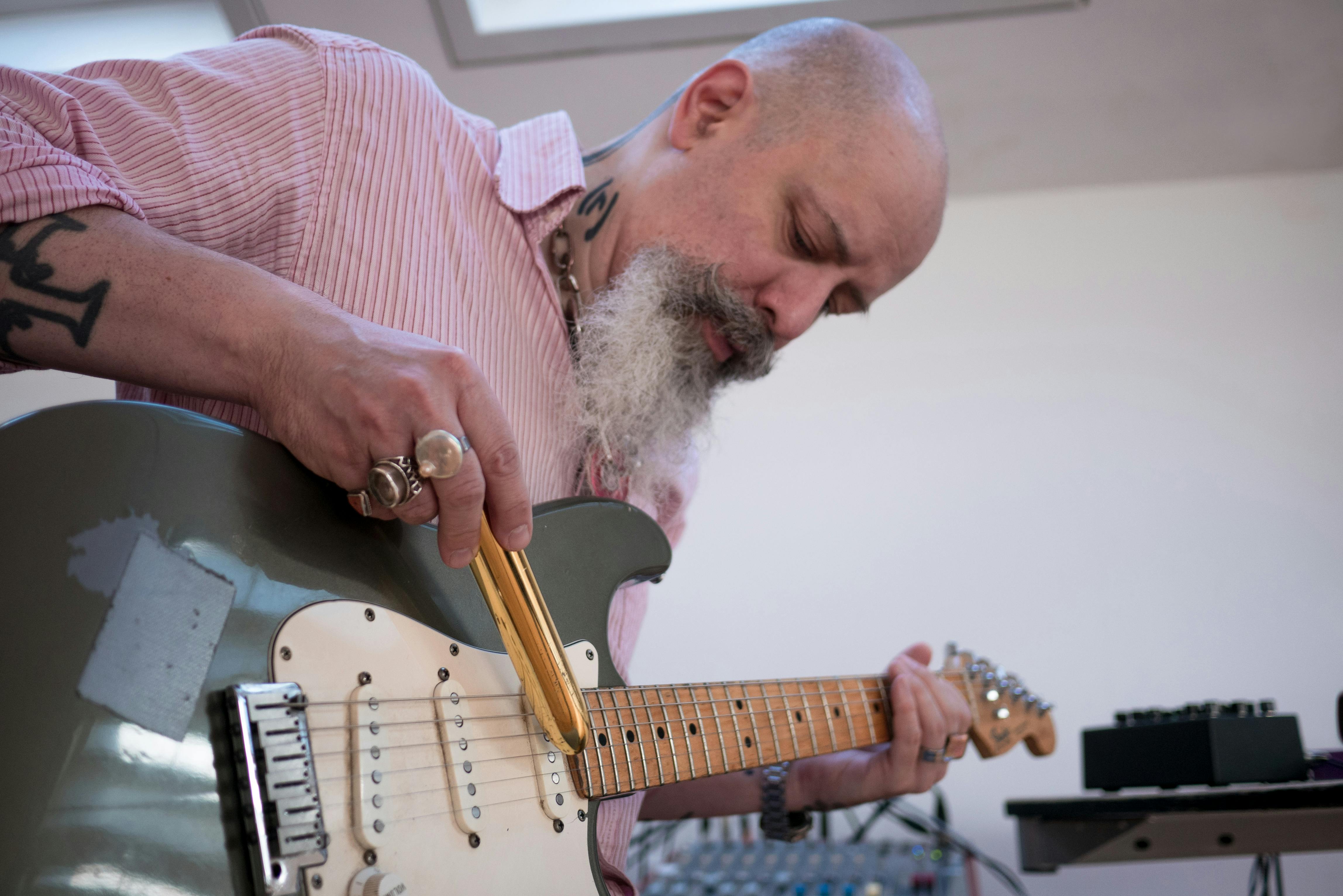 Il musicista Fabrizio Modonese Palumbo mentre suona la chitarra elettrica. Il viso concentrato è incorniciato da una lunga barba bianca; indossa una camicia rosa a righe e numerosi anelli. Su braccia e collo si intravedono dei tatuaggi.