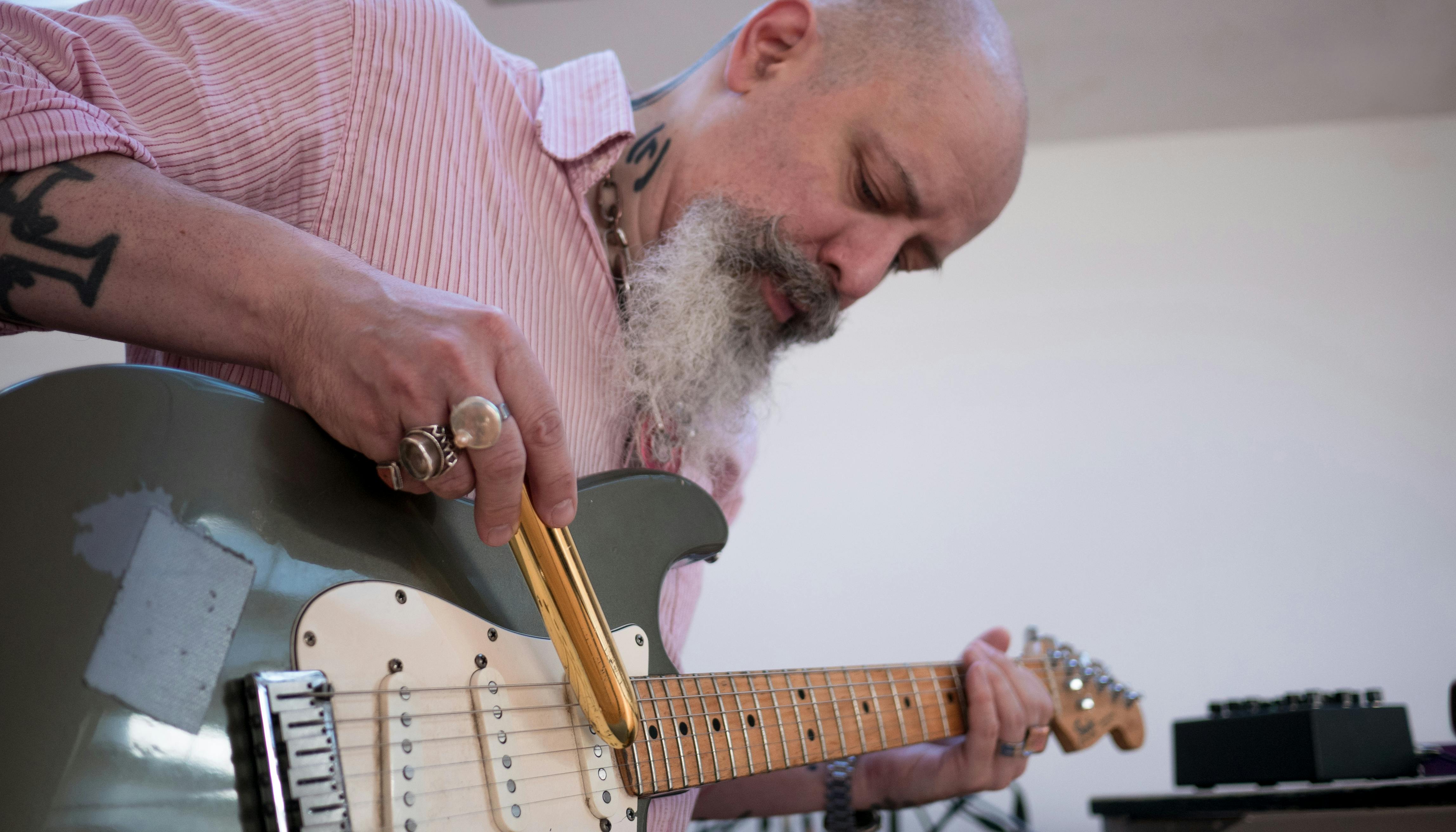 Musician Fabrizio Modonese Palumbo as he plays electric guitar. His concentrated face is framed by a long white beard; he wears a pink striped shirt and numerous rings. Tattoos can be glimpsed on his arms and neck.
