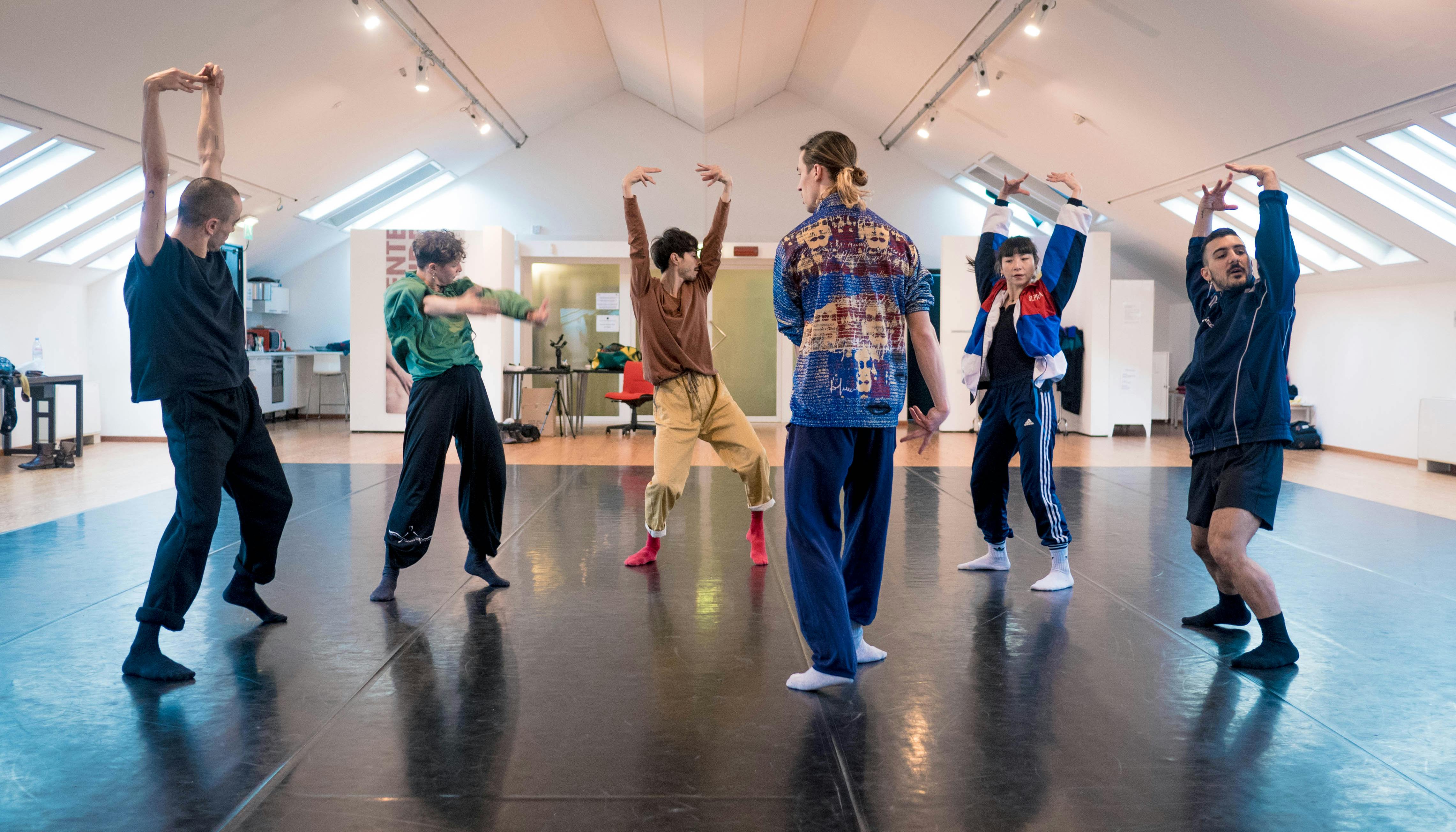 Six dancers are rehearsing in the Studio. They are standing in a circle, facing each other. They wear colorful jumpsuits and socks.