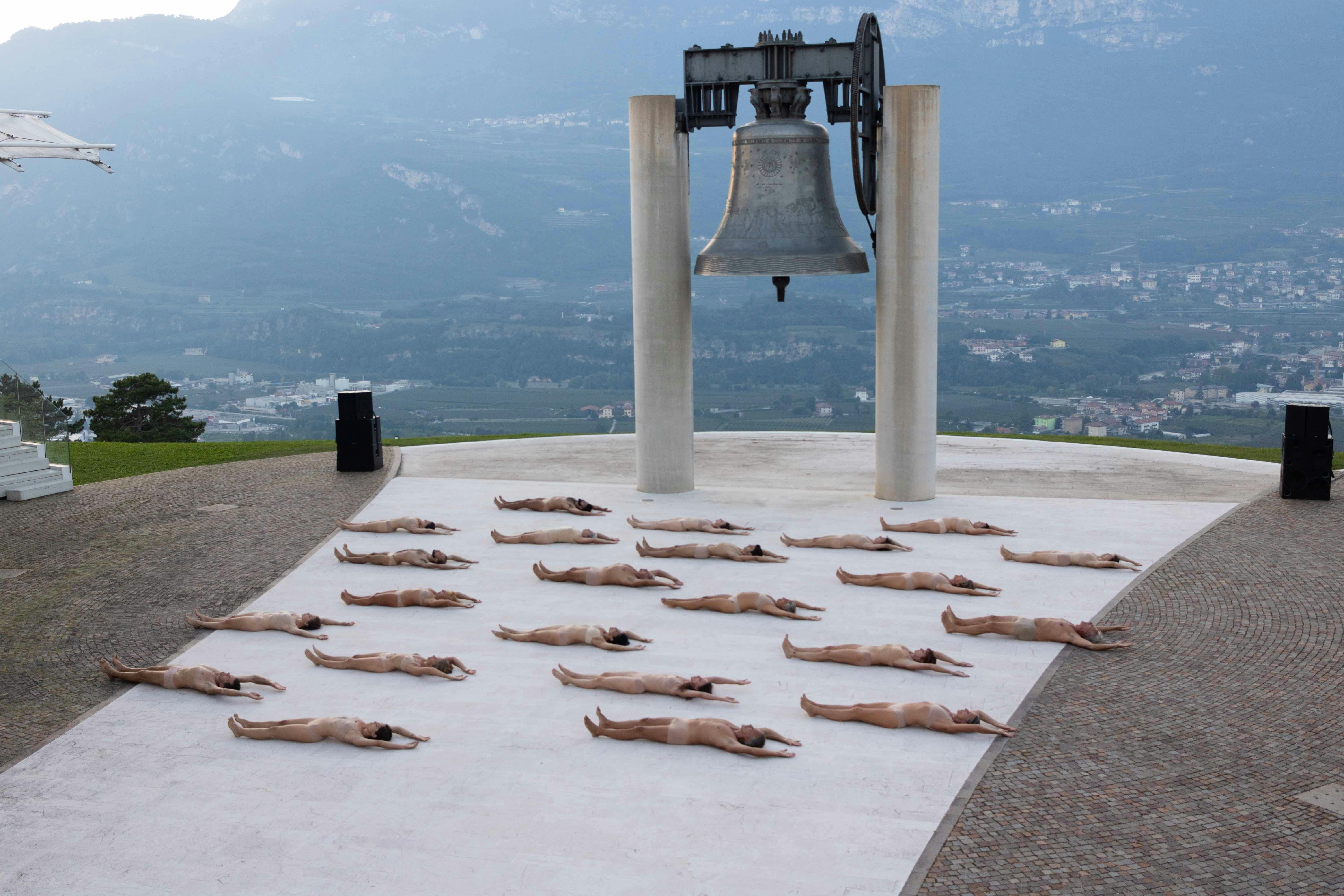In front of the Campana dei Caduti in Rovereto, numerous human bodies are lying on the ground, facing upwards, naked. Their arrangement in space is orderly.