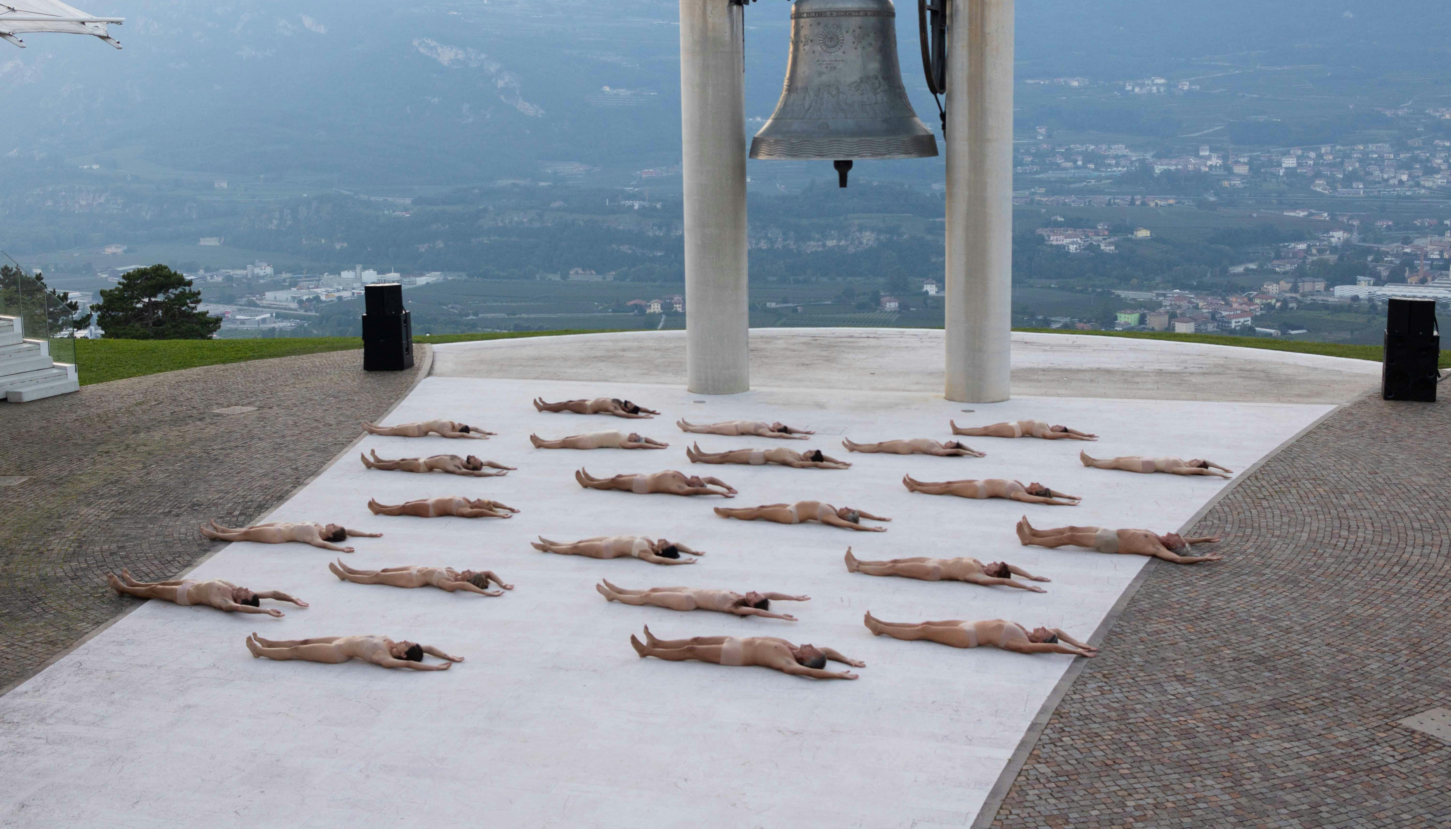 Alla base della Campana dei Caduti di Rovereto, numerosi corpi umani sono sdraiati a terra, rivolti verso l'alto, nudi. La loro disposizione nello spazio è ordinata.