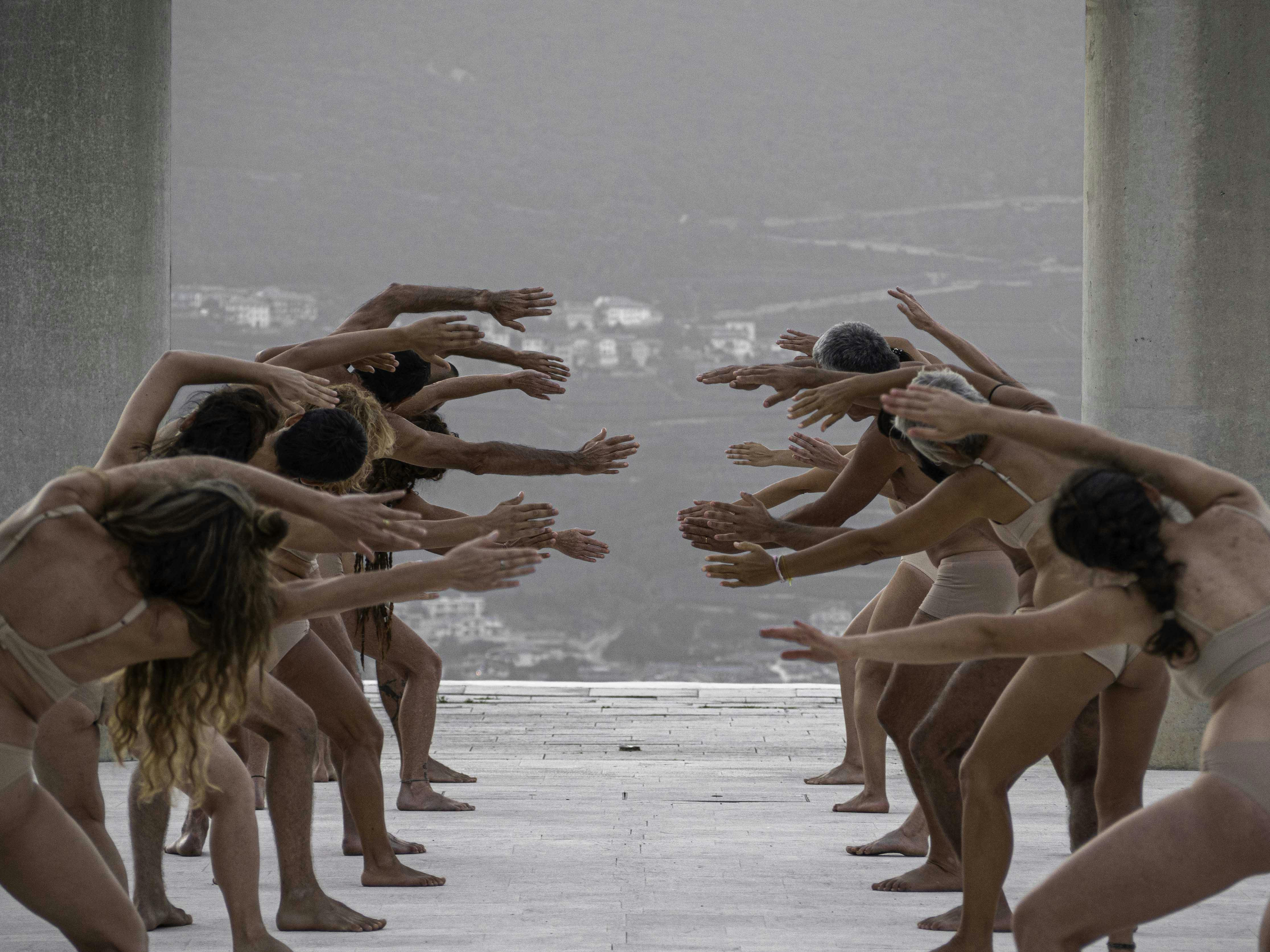 Two parallel rows of human bodies, shot frontally. The performers, wearing only flesh-colored underwear, stretch their arms and torso toward the center, trying to push themselves toward the opposite row.