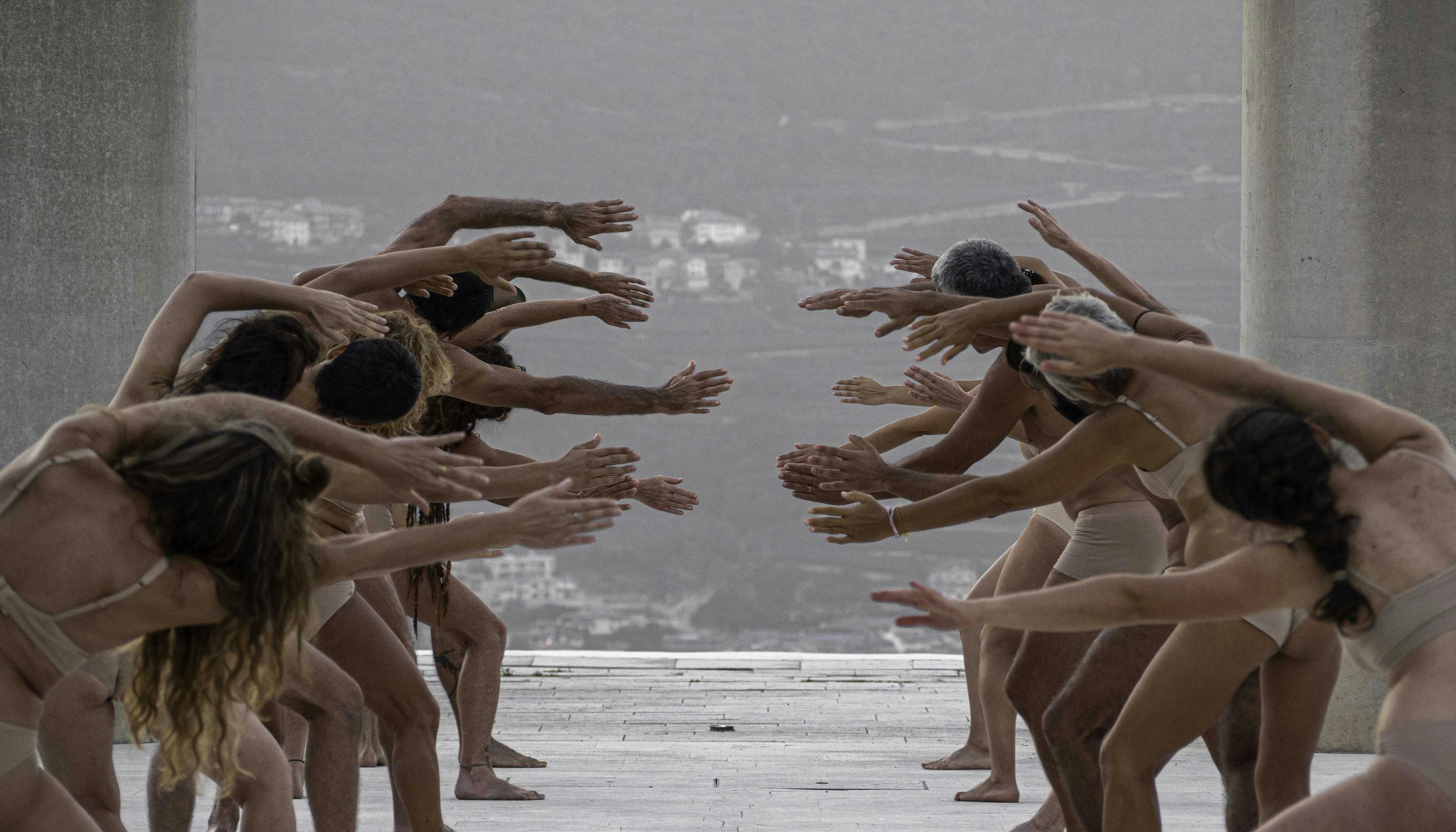 Two parallel rows of human bodies, shot frontally. The performers, wearing only flesh-colored underwear, stretch their arms and torso toward the center, trying to push themselves toward the opposite row.