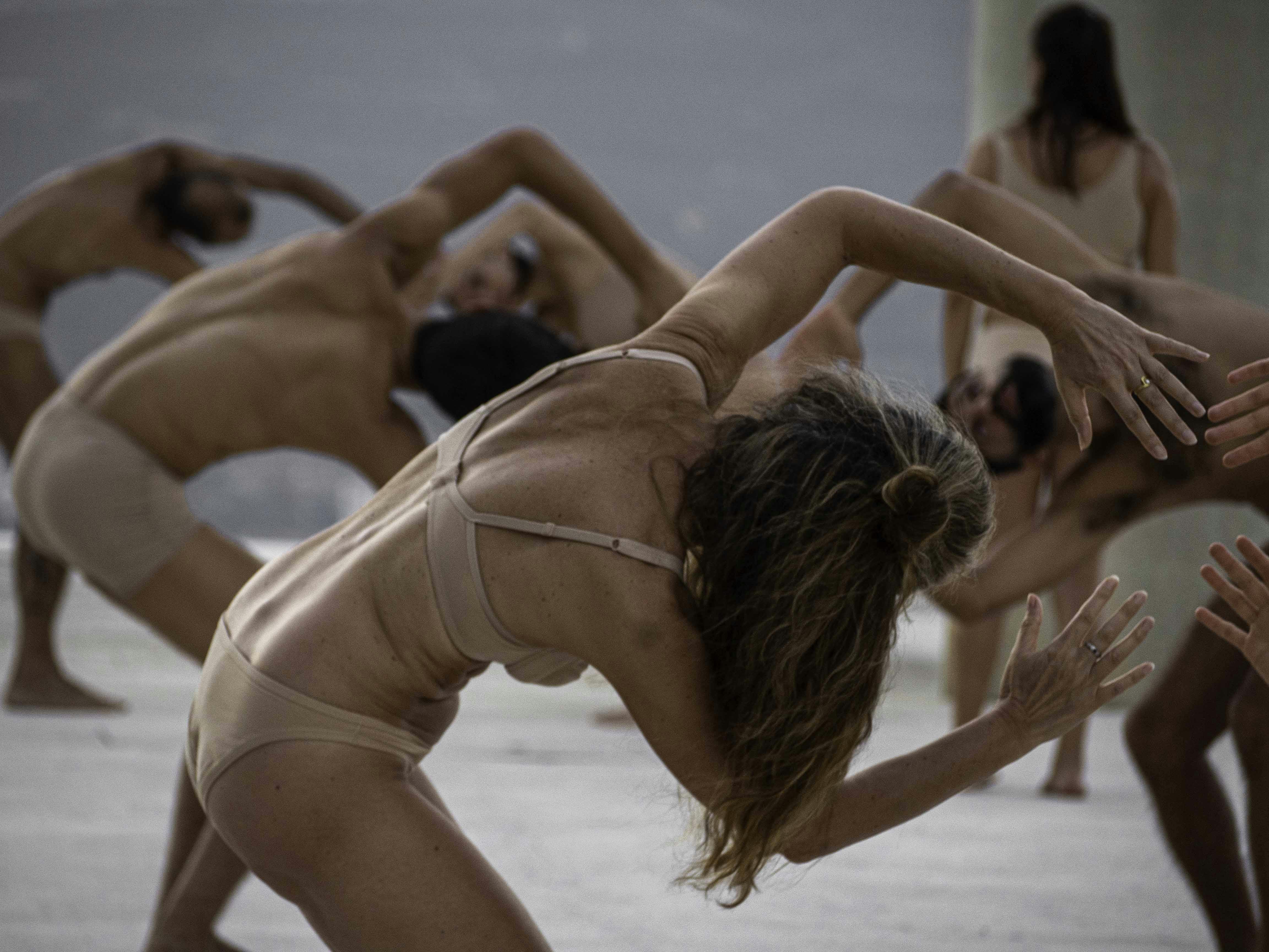Several performers are seen with their backs to the camera, leaning forward with their torso and arms in a circle above their heads. They wear only flesh-colored underwear. The performance takes place outdoors.