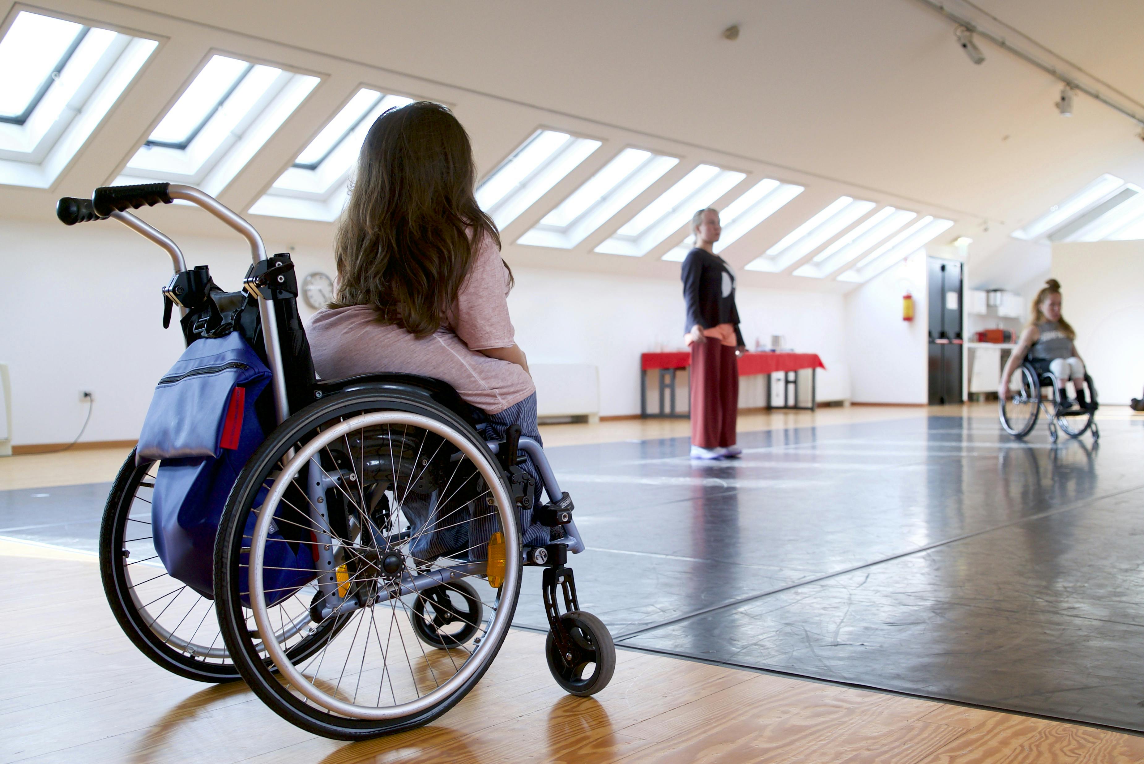 In the foreground Chiara Bersani, on her back, in a wheelchair, observes the dancers of Danskomapaniet SPINN moving through space. In the distance we see a dancer standing and a second one sitting in a wheelchair.