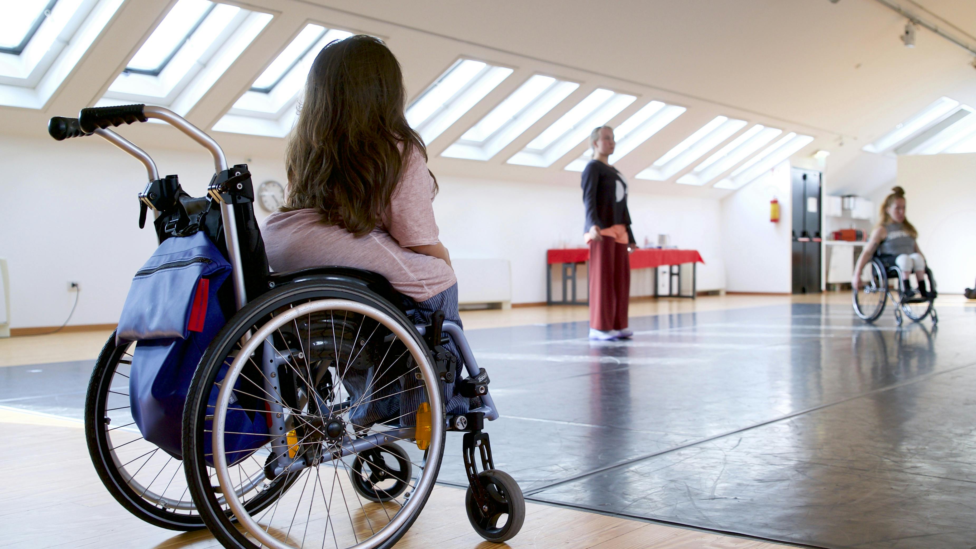 In the foreground Chiara Bersani, on her back, in a wheelchair, observes the dancers of Danskomapaniet SPINN moving through space. In the distance we see a dancer standing and a second one sitting in a wheelchair.