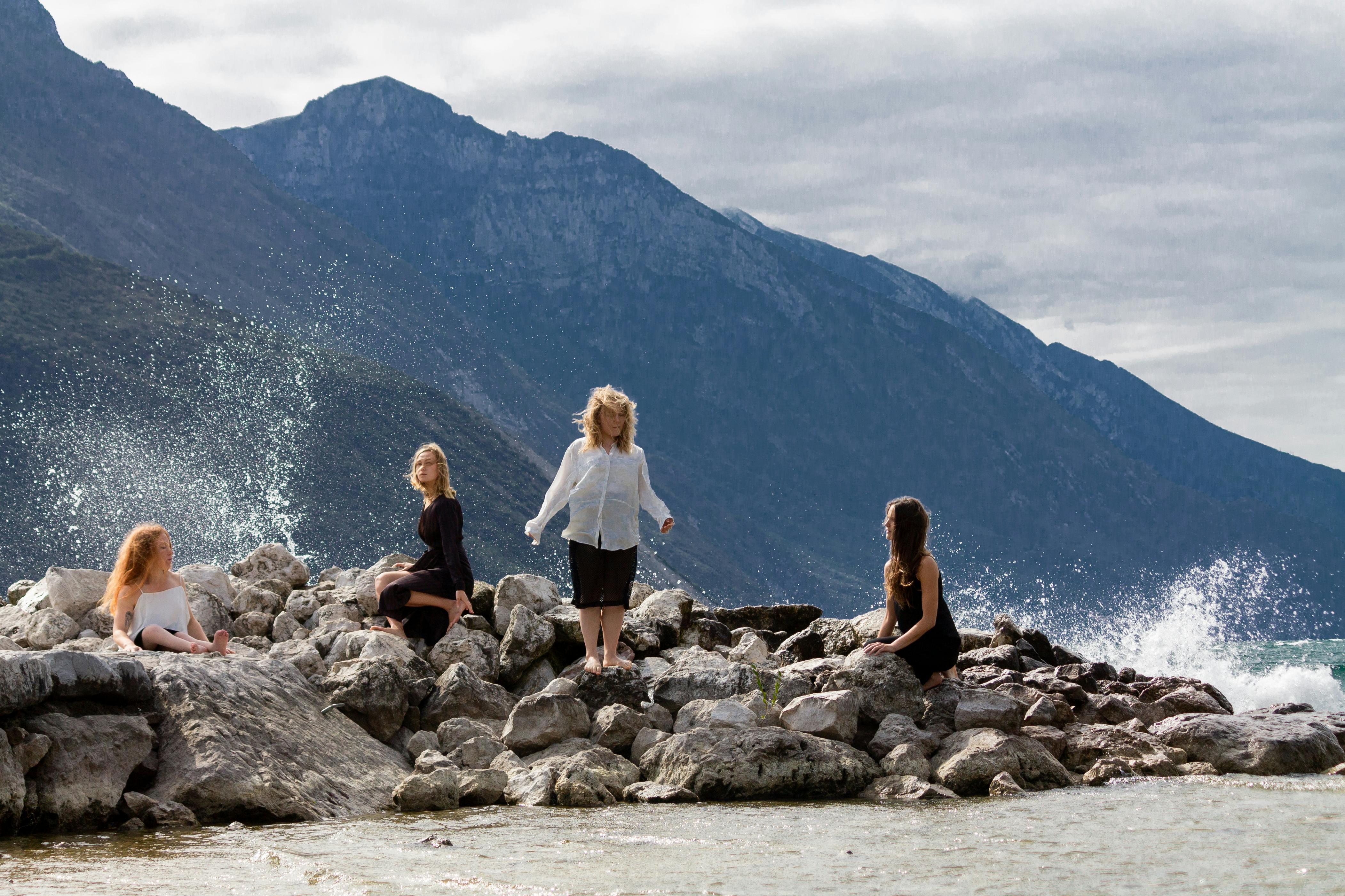 Cliffs can be seen on a water surface; mountains in the background.  Four women are standing or sitting on the rocks, standing still and waiting. Splashes of water frame their figures.