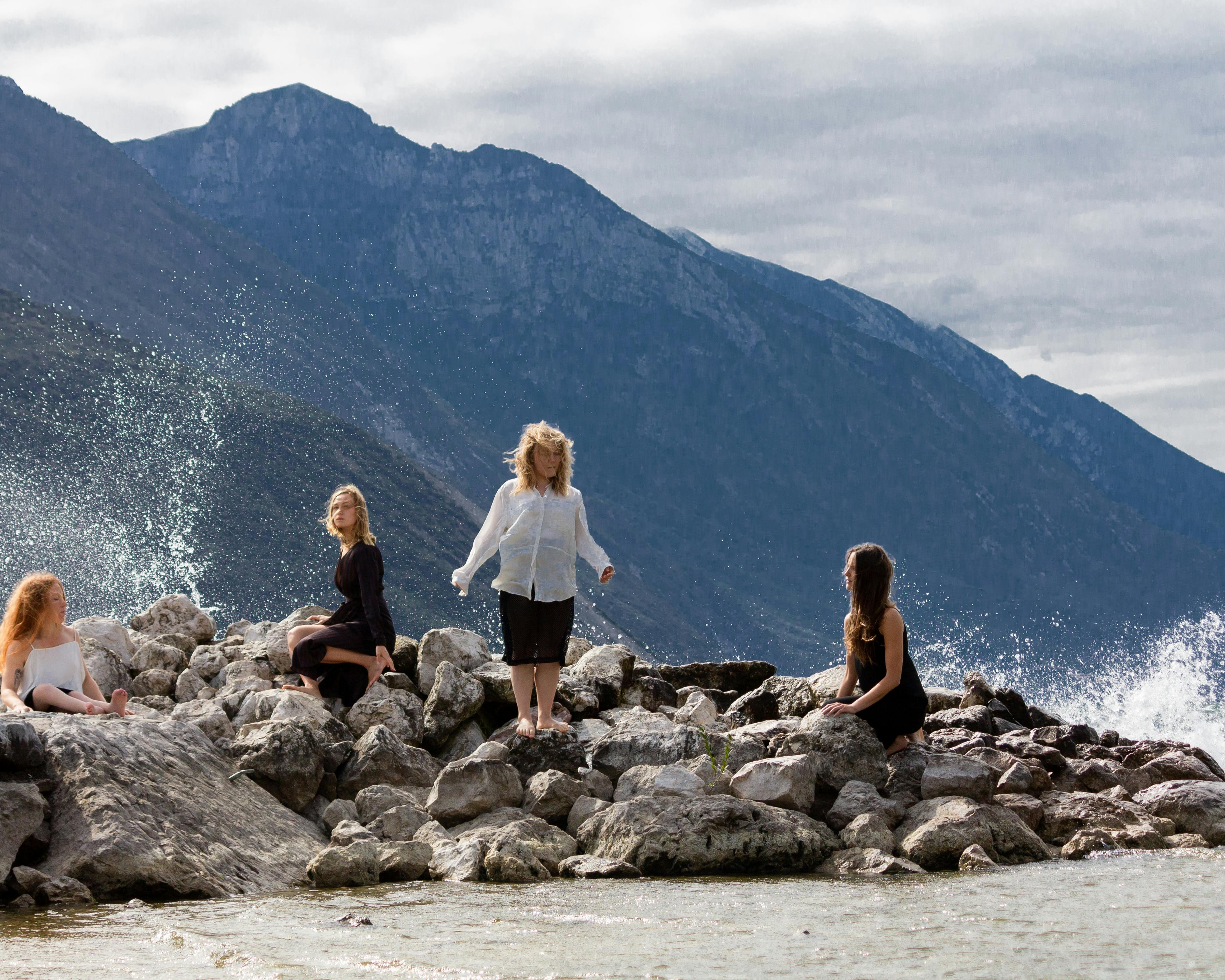 Si vedono delle rocce su una superficie d'acqua; sullo sfondo le montagne.  Quattro donne sono in piedi o sedute sulle rocce, ferme in attesa. Spruzzi d'acqua ne incorniciano le figure.