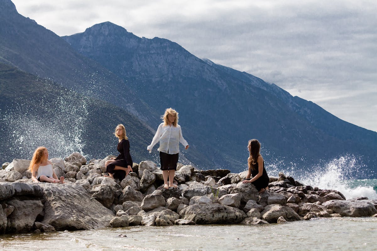 Cliffs can be seen on a water surface; mountains in the background.  Four women are standing or sitting on the rocks, standing still and waiting. Splashes of water frame their figures.