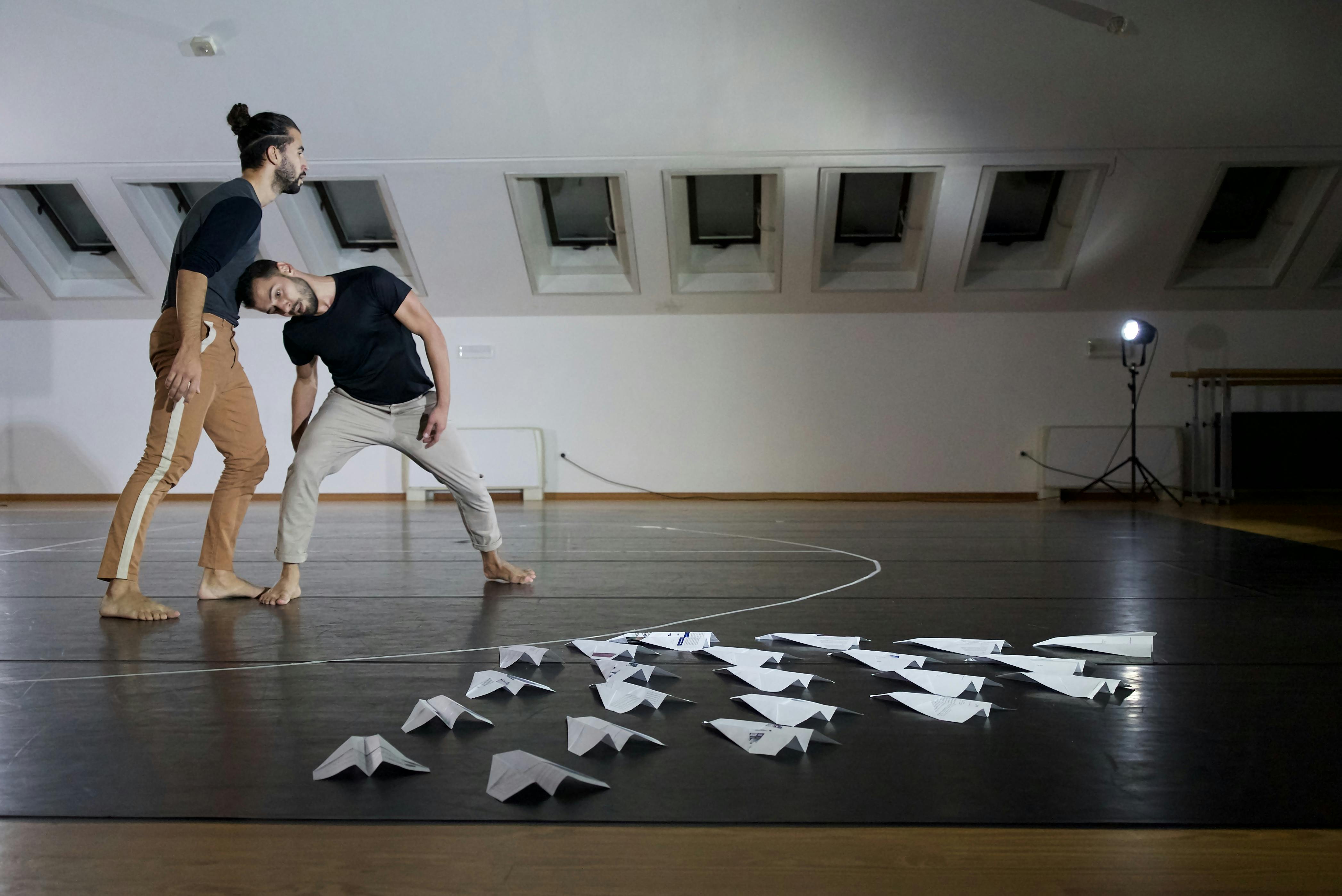 The two choreographers and dancers rehearse in the room: one of them puts his weight on the head of the other, leaning to support him. On the floor there are several paper airplanes arranged in a triangle. 
