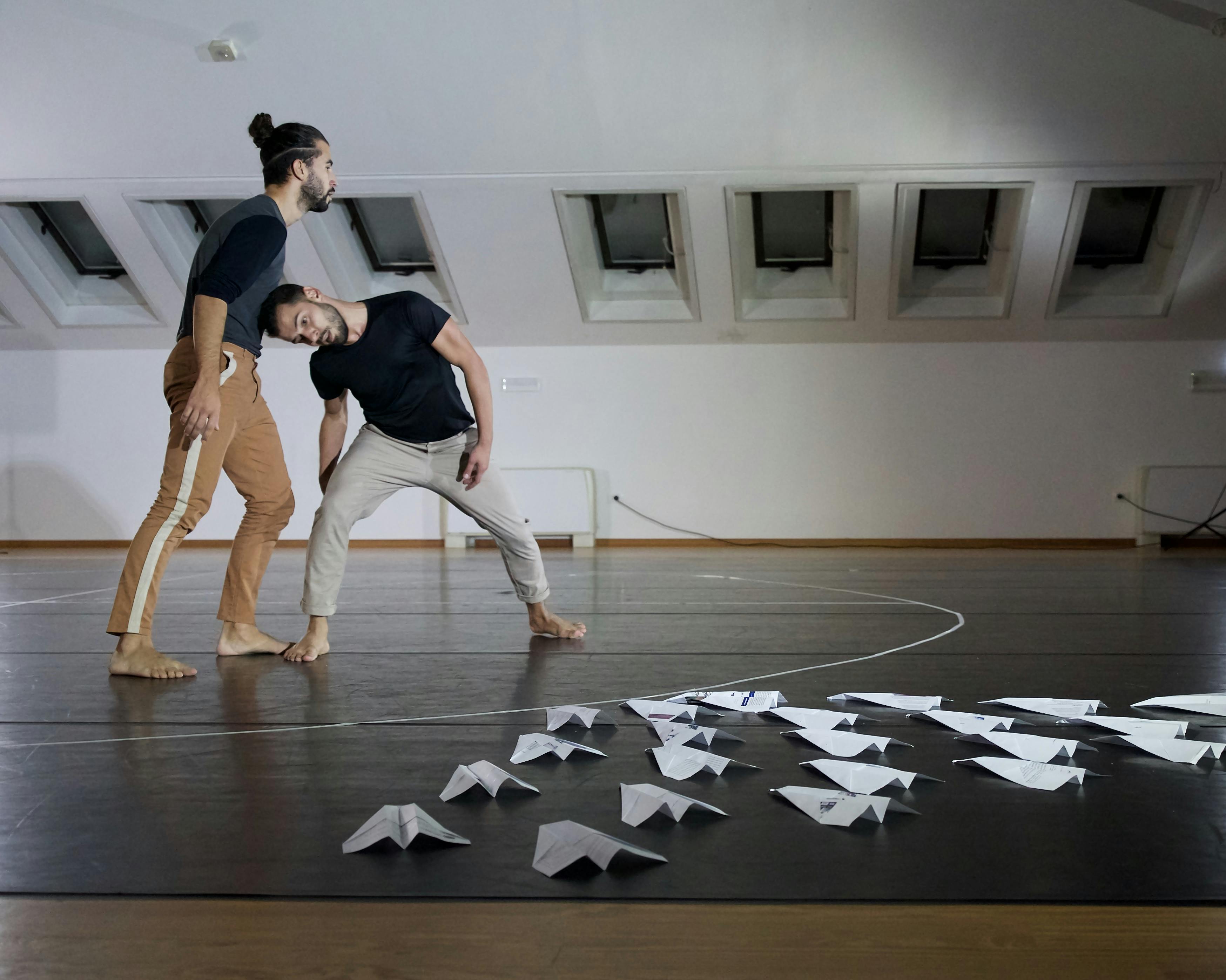 I due coreografi e danzatori provano in sala: uno dei due appoggia il proprio peso sulla testa dell'altro, piegato per sostenerlo. Per terra ci sono numerosi areoplanini di carta disposti a triangolo. 