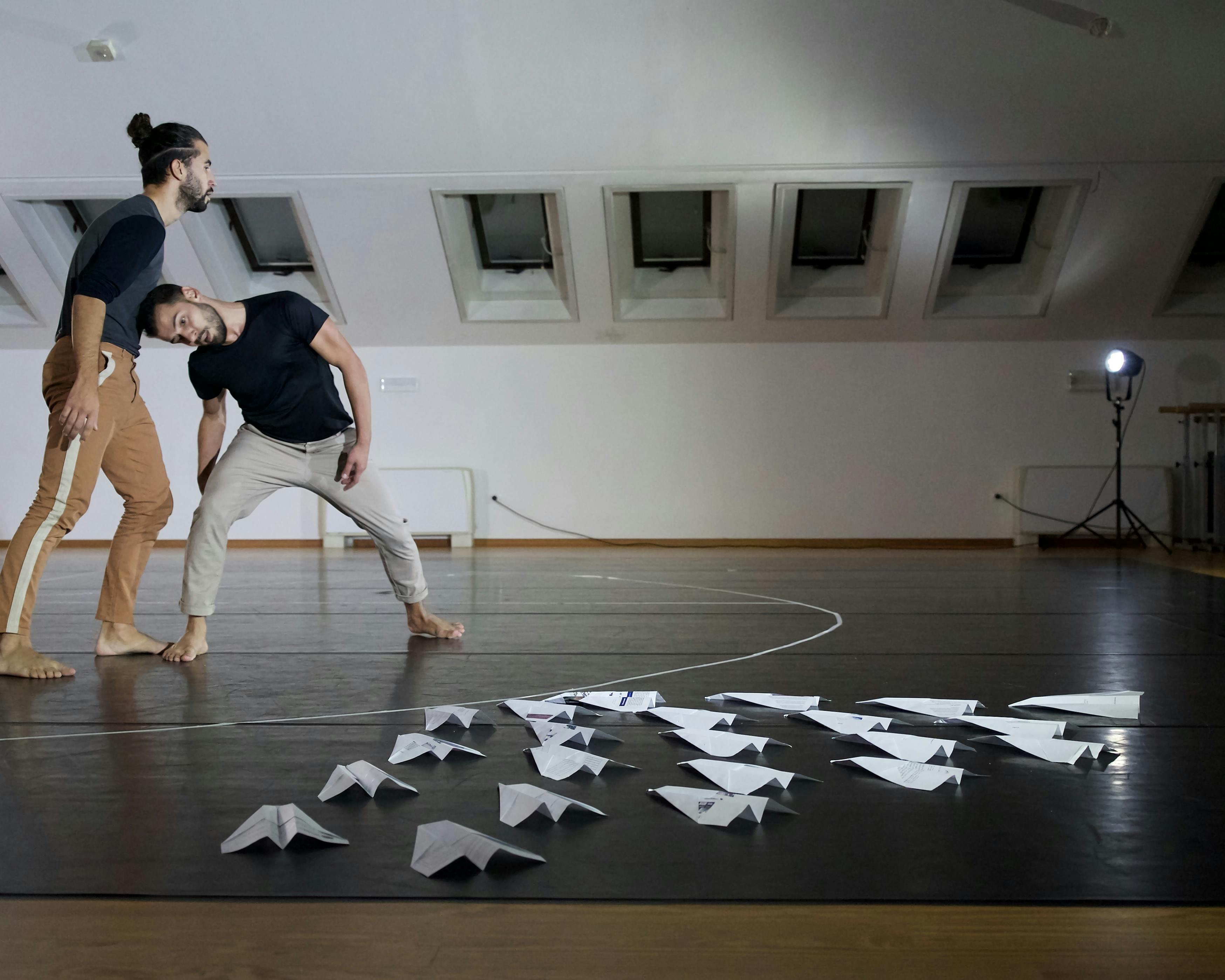 The two choreographers and dancers rehearse in the room: one of them puts his weight on the head of the other, leaning to support him. On the floor there are several paper airplanes arranged in a triangle. 