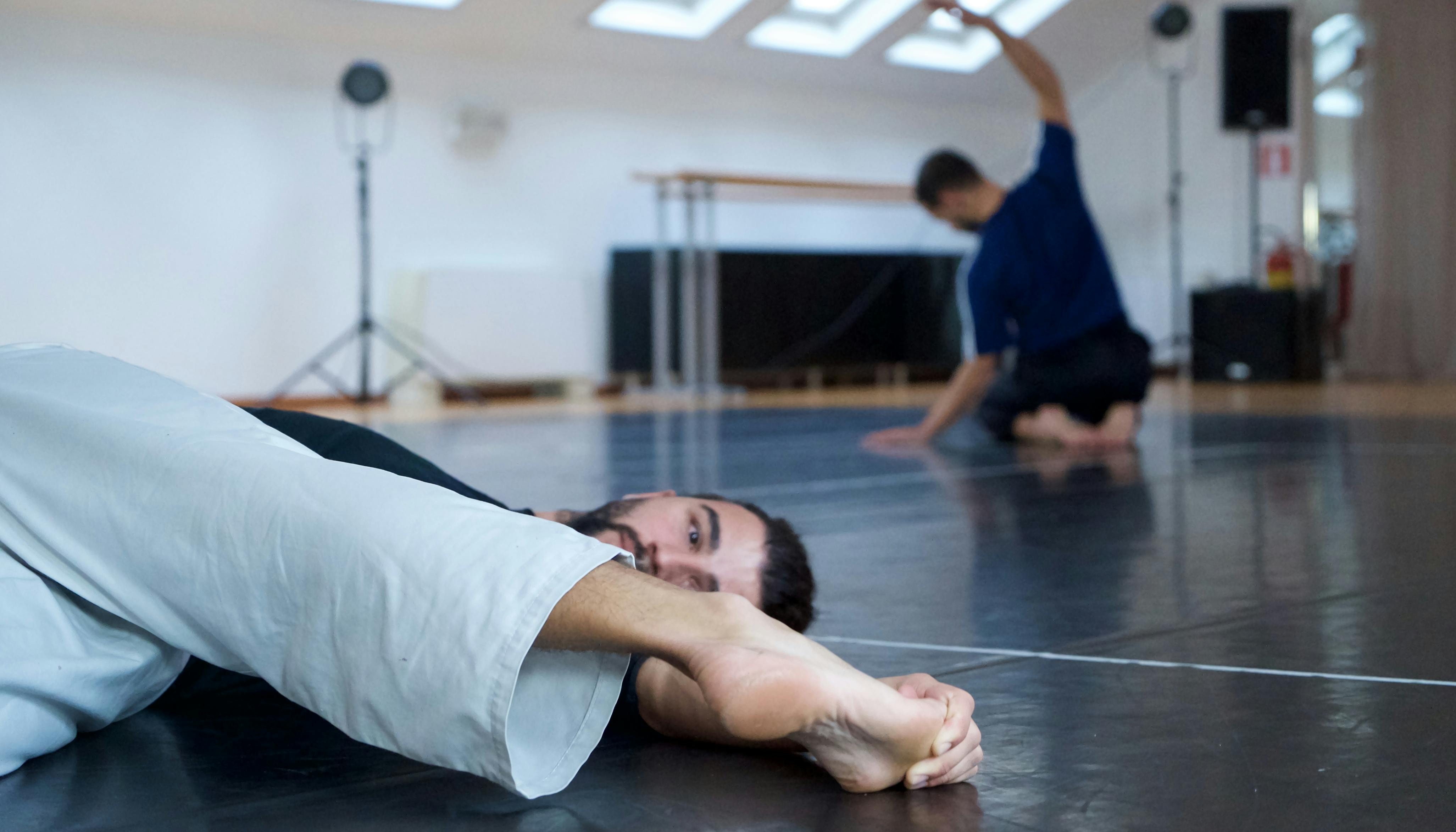 The two choreographers warm up before rehearsal: one of them is lying on the floor, the other on his knees stretching his back.