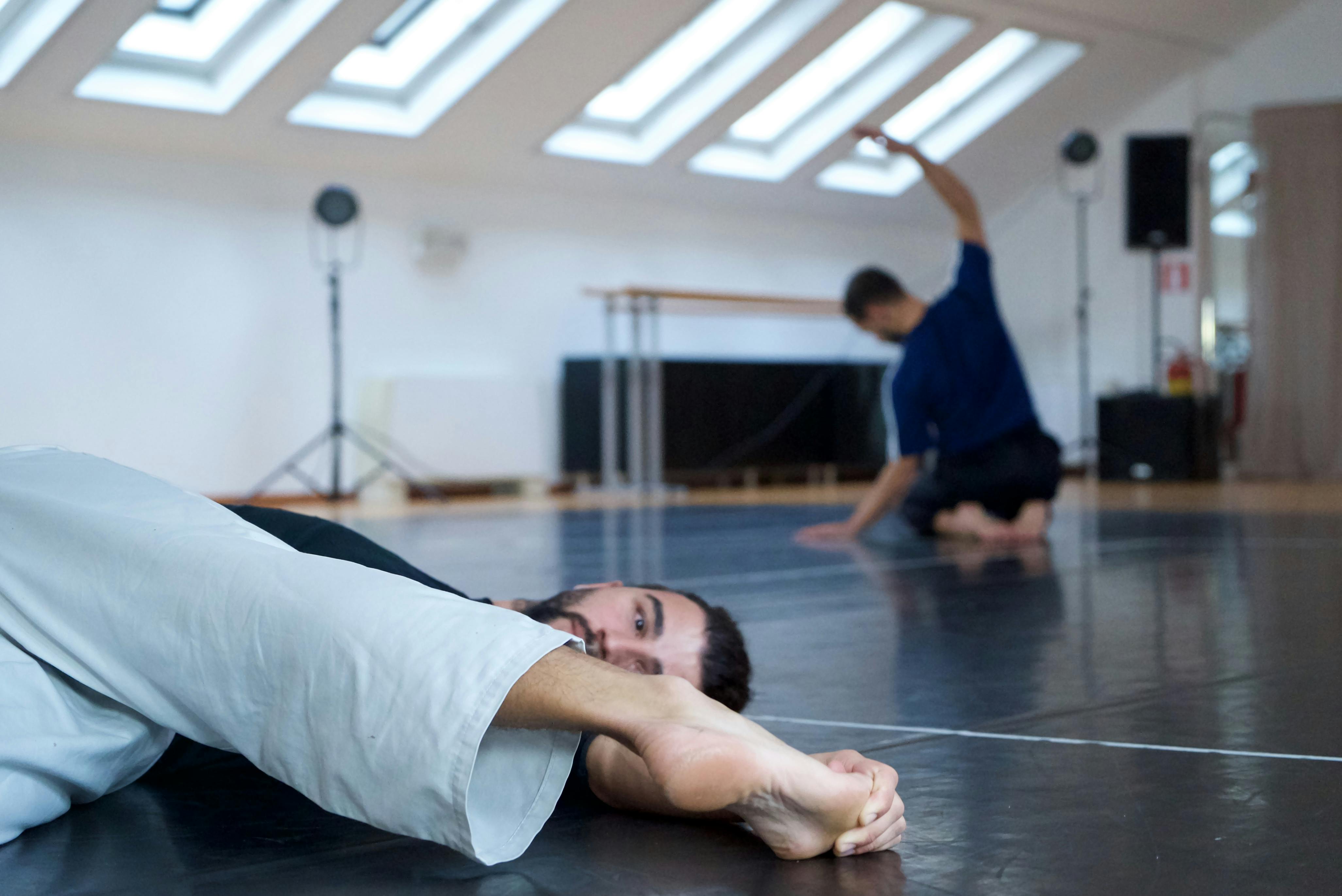 I due coreografi si riscaldano prima delle prove: uno dei due è sdraiato a terra, l'altro in ginocchio allunga la schiena.