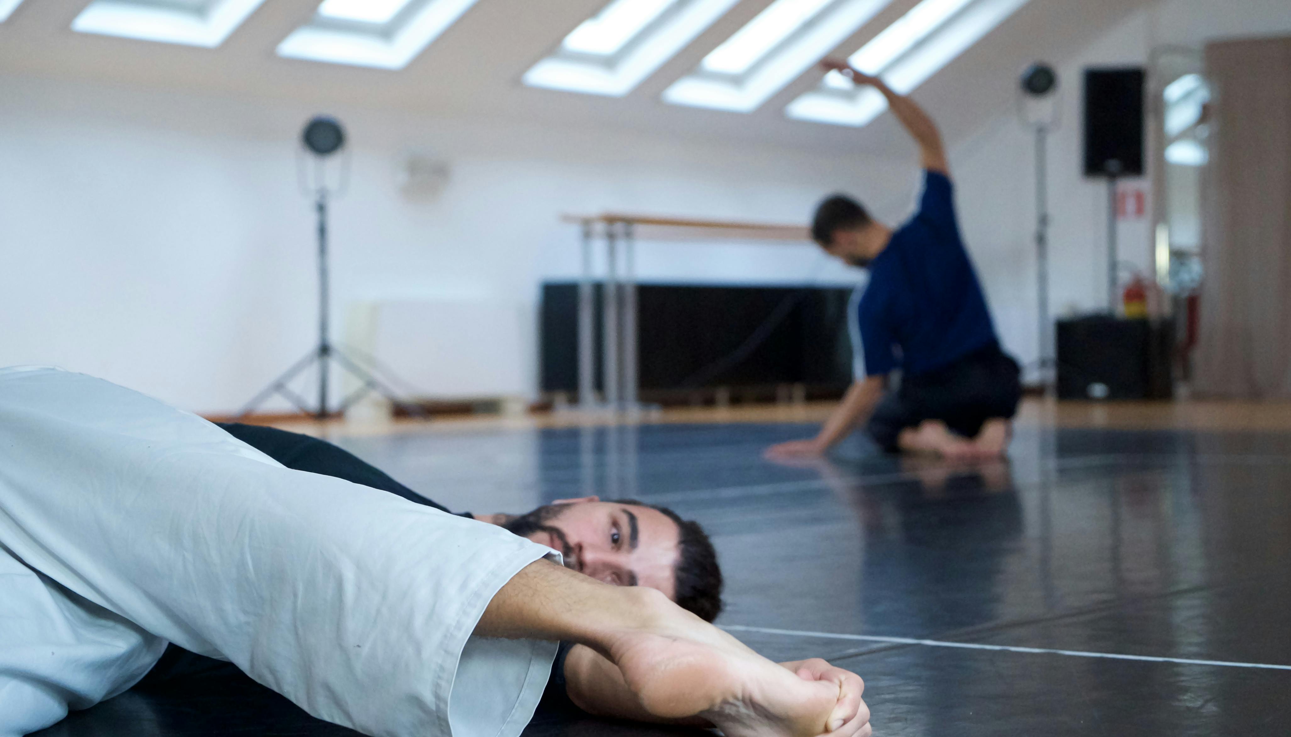 The two choreographers warm up before rehearsal: one of them is lying on the floor, the other on his knees stretching his back.