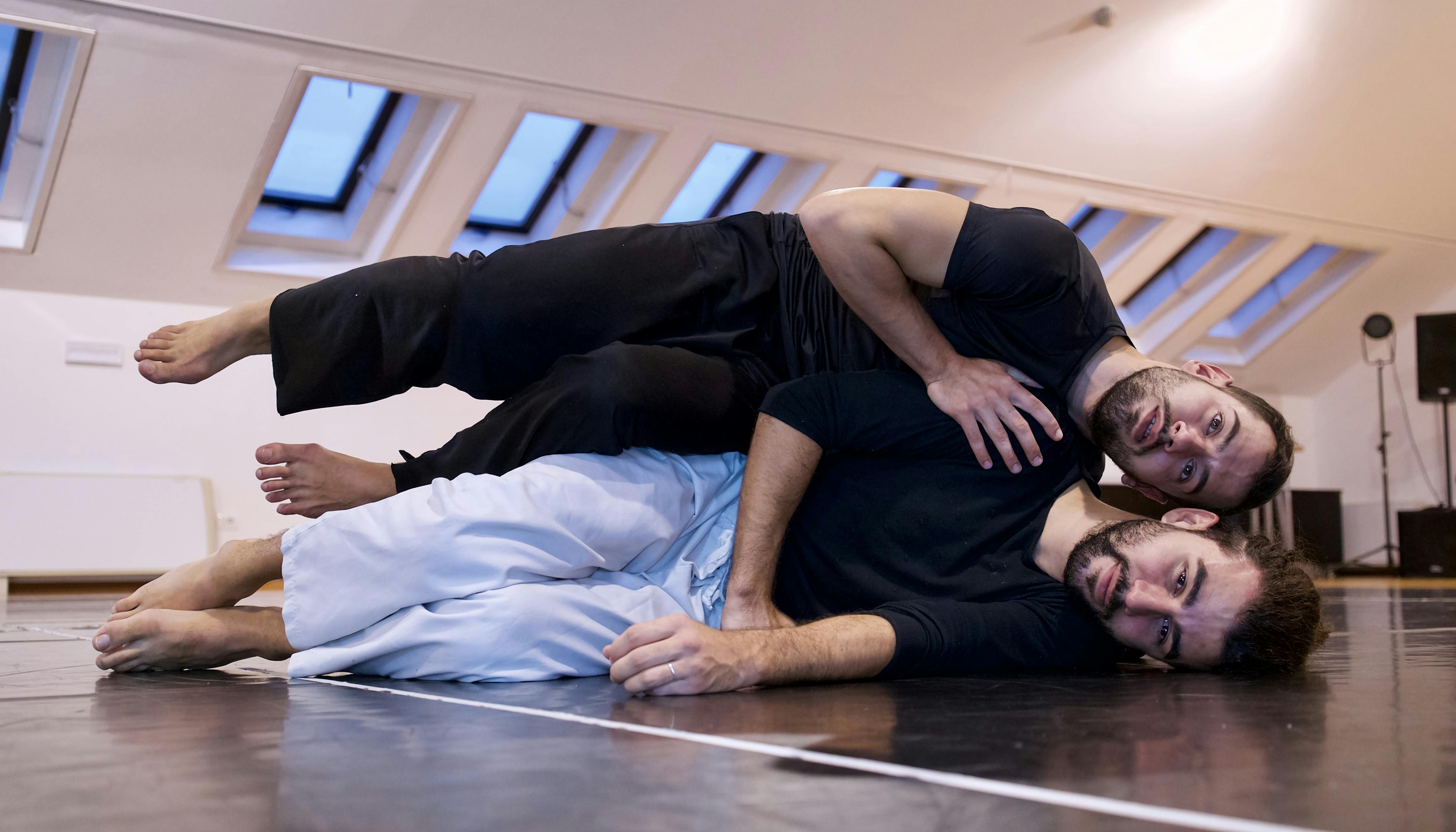 The two dancers are stretched out on the floor on one side, facing the camera, one suspended over the other's body.