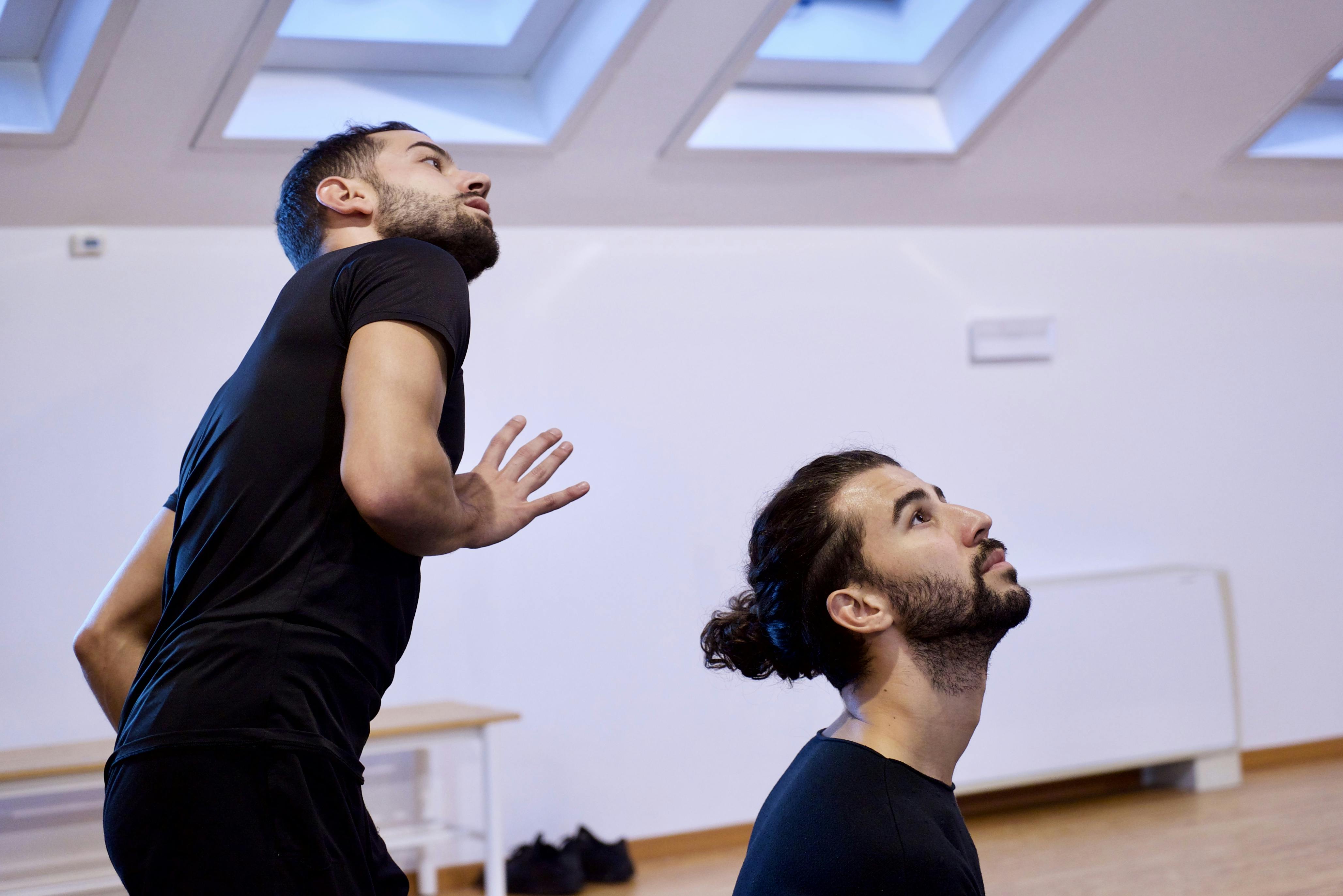 Prove in sala. I due danzatori hanno lo sguardo rivolto leggermente verso l'alto. Sono in tuta, di uno vediamo tutta la parte superiore del corpo, dell'altro solo testa e spalle.