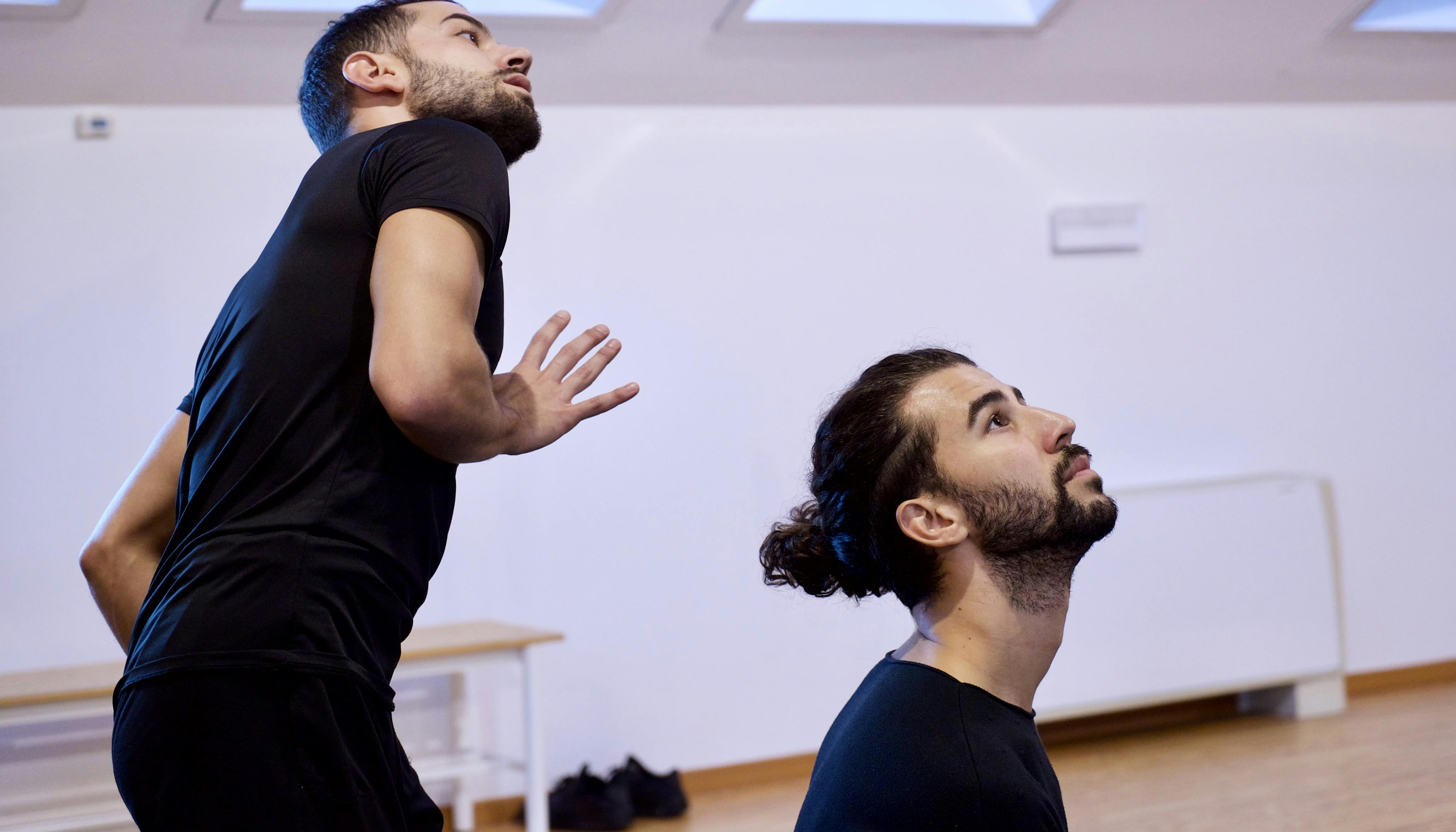 Rehearsal in the Studio. The two dancers are looking slightly upwards. They are in suits, we can see the whole upper part of one's body, only the head and shoulders of the other.