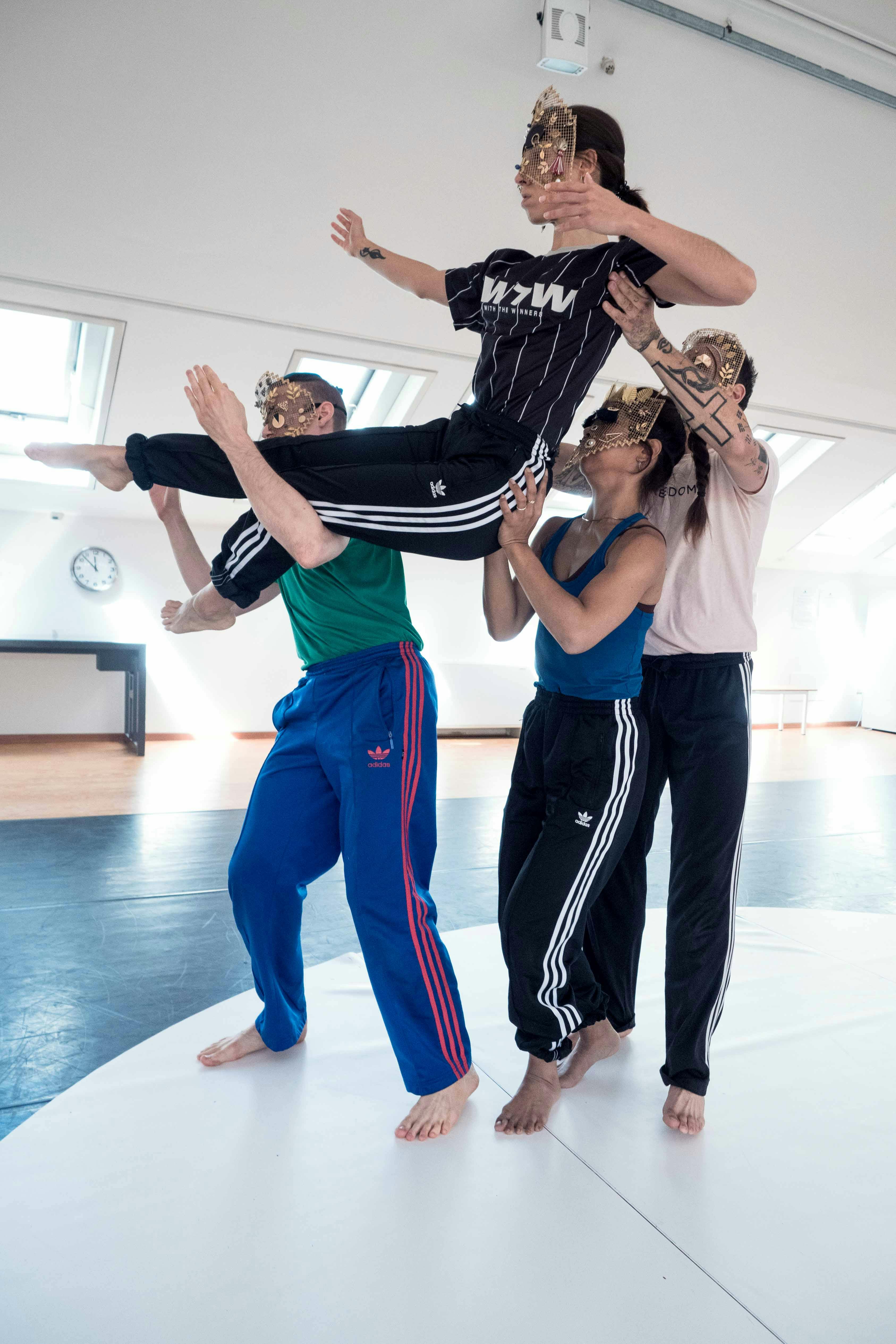 A dancer is lifted by three performers; she is in a seated position with her legs stretched forward. Everyone's arms are in a rigid position.