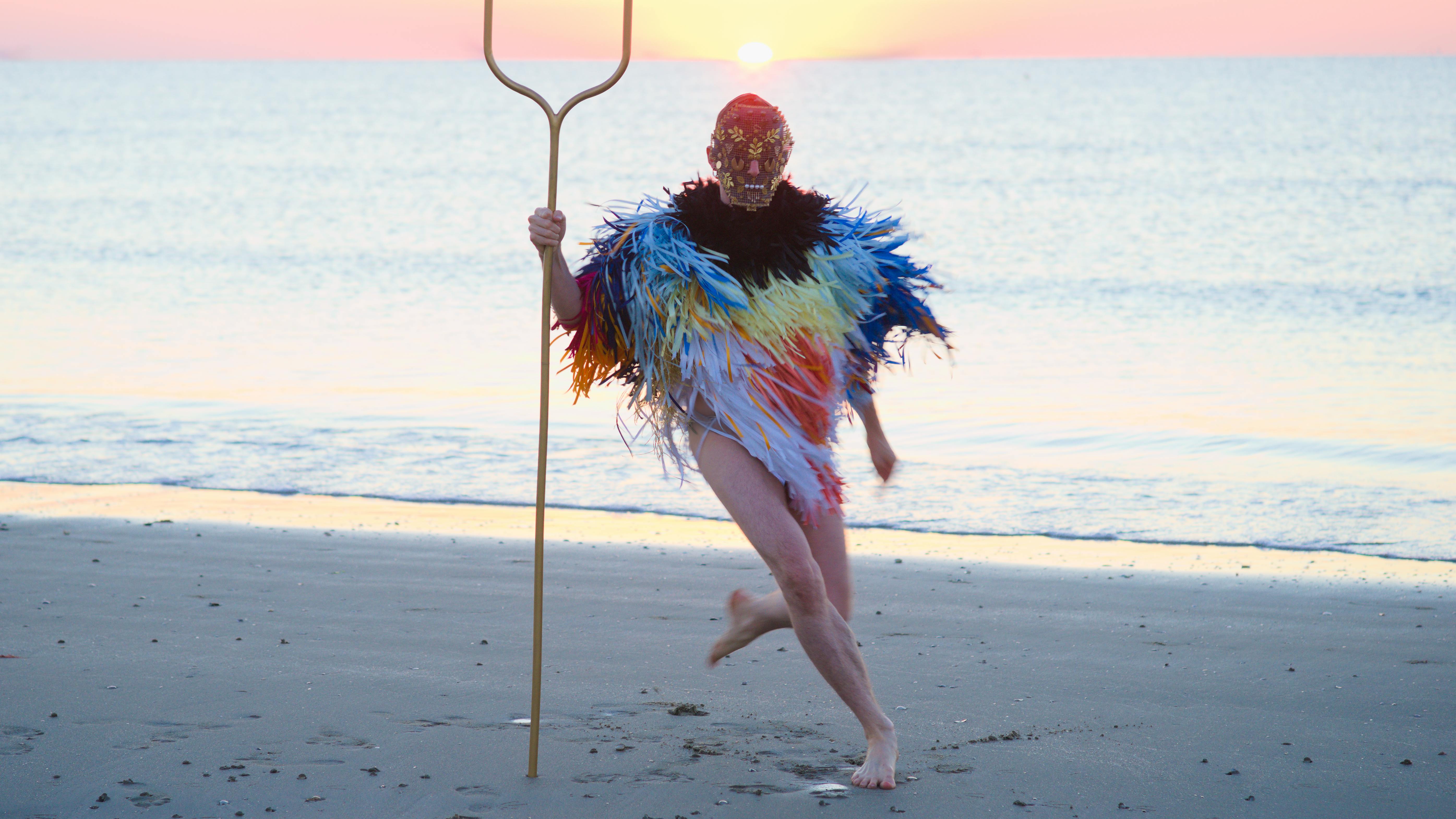 Una figura umana, con indosso una maschera e un poncho fatto di sottili striscioline colorate, corre su una spiaggia. In mano tiene una specie di forcone  a due punte.