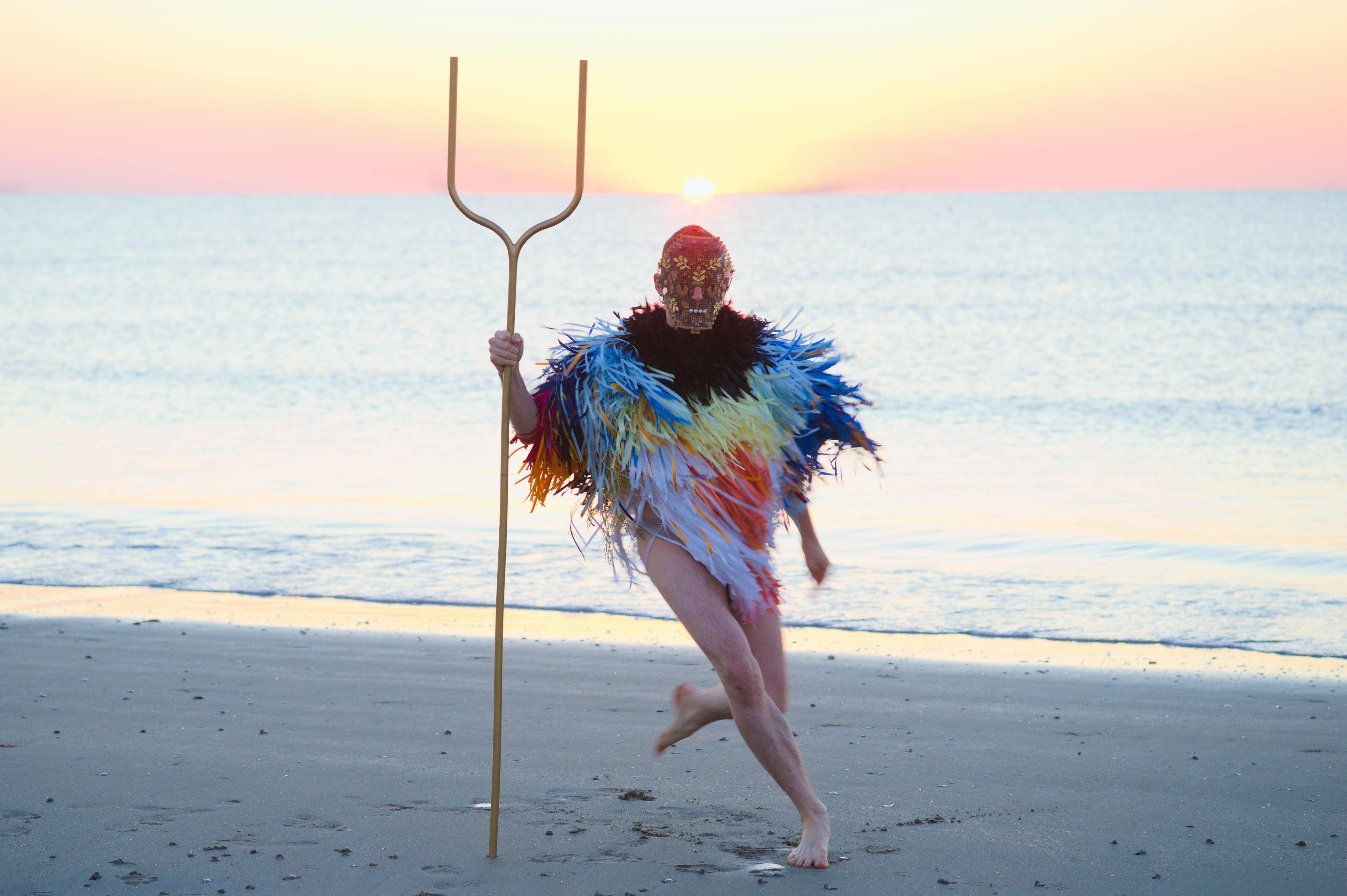 A human figure, wearing a mask and a poncho made of thin colored strips, is running on a beach. In their hand they holds a kind of two-pronged pitchfork.