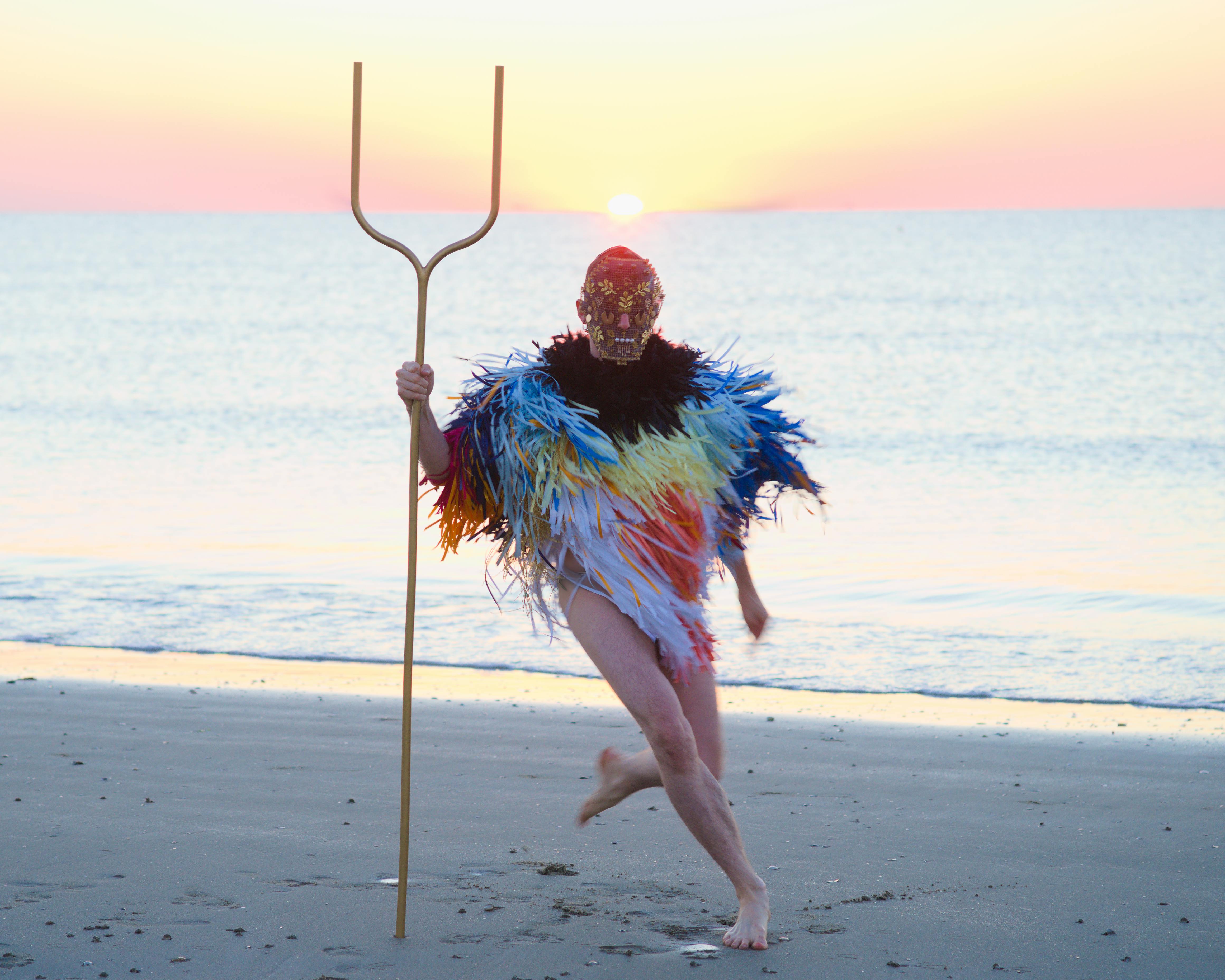A human figure, wearing a mask and a poncho made of thin colored strips, is running on a beach. In their hand they holds a kind of two-pronged pitchfork.
