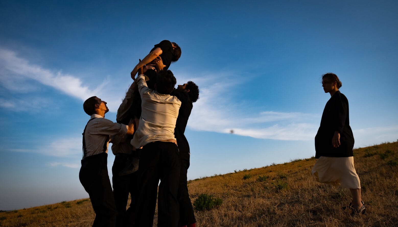 Three men lift a woman vertically up a slope. The two behind support her by the shoulders, while the one in front holds her by the legs. The woman is at dead weight, while a second one looks at the scene on the right.