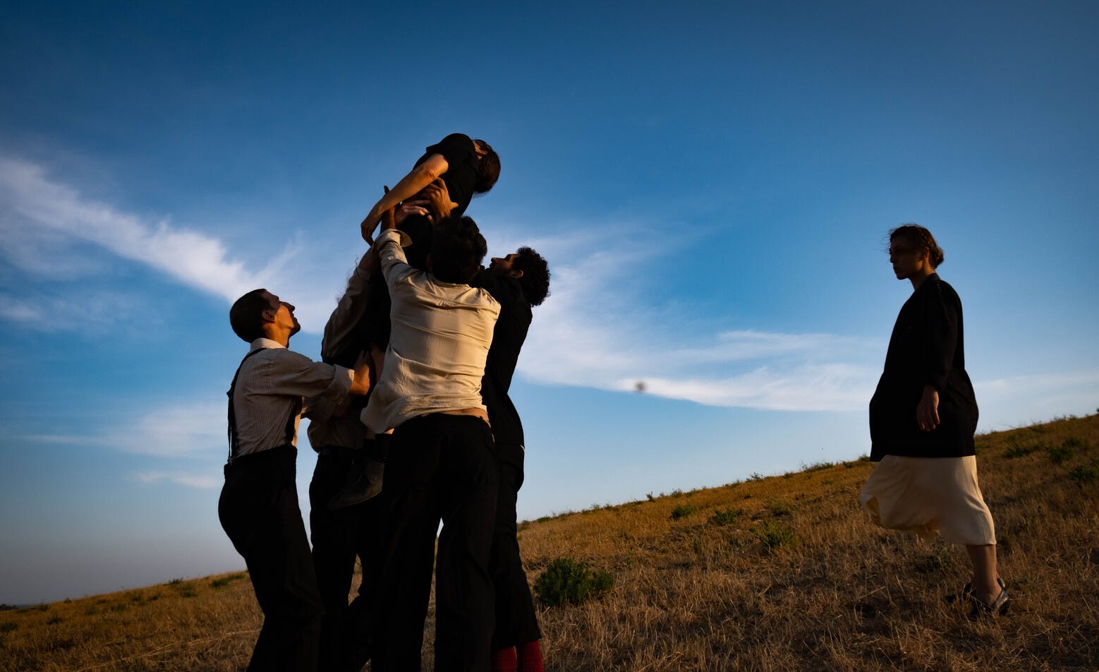 Three men lift a woman vertically up a slope. The two behind support her by the shoulders, while the one in front holds her by the legs. The woman is at dead weight, while a second one looks at the scene on the right.