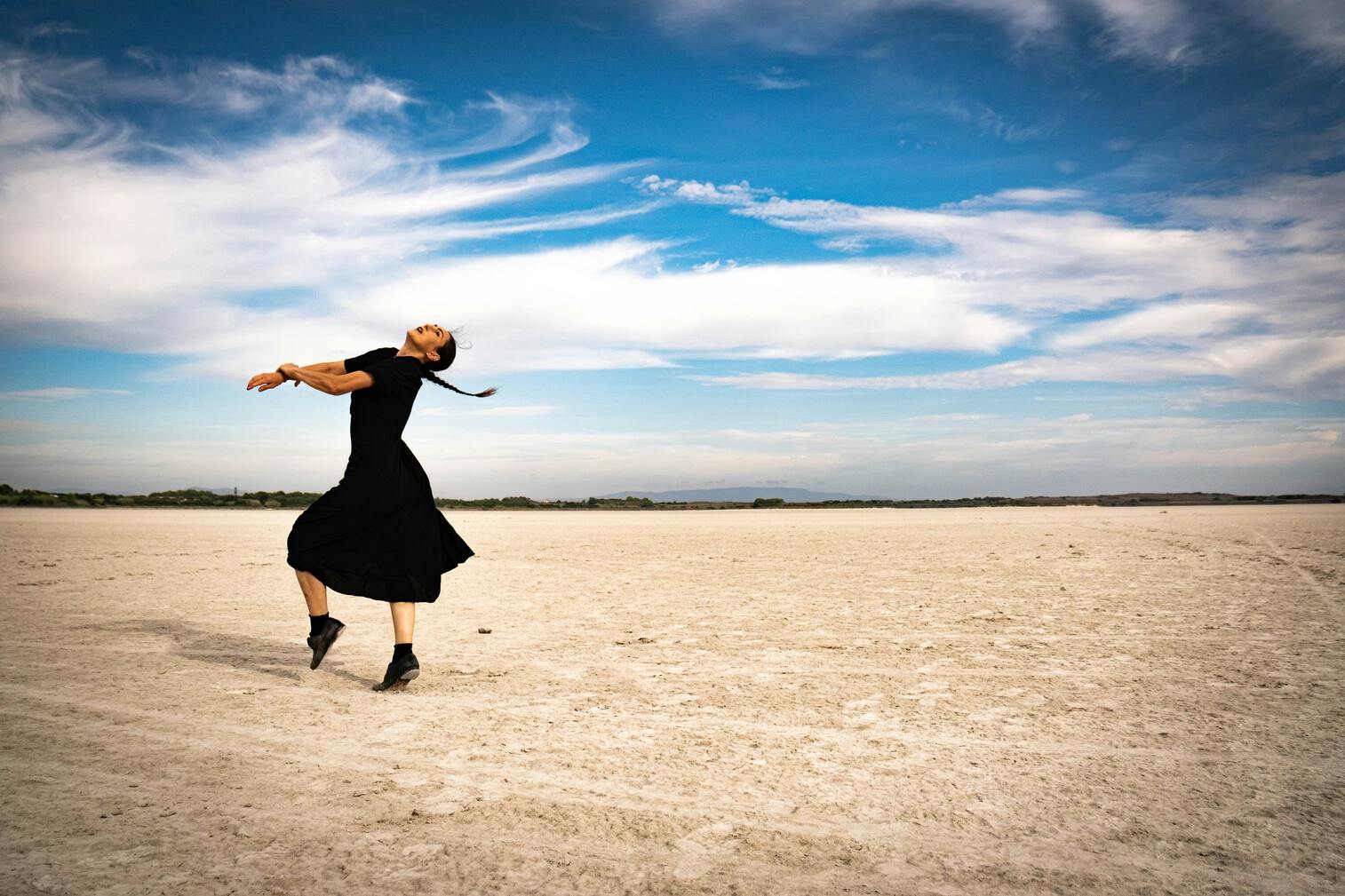 A dancer is dancing on a beach. She stretches her arms forward while extending her head back.