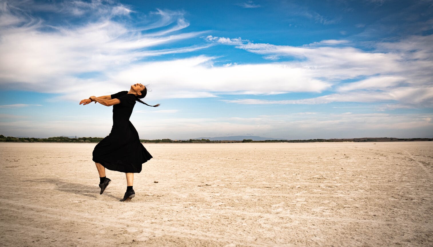 Una danzatrice sta danzando su di una spiaggia. Protende le braccia in avanti mentre distende la testa indietro.