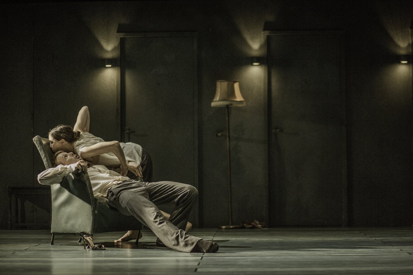 A man, lying on an armchair, stretches his legs forward. A woman, standing to the side, stretches her torso over him.