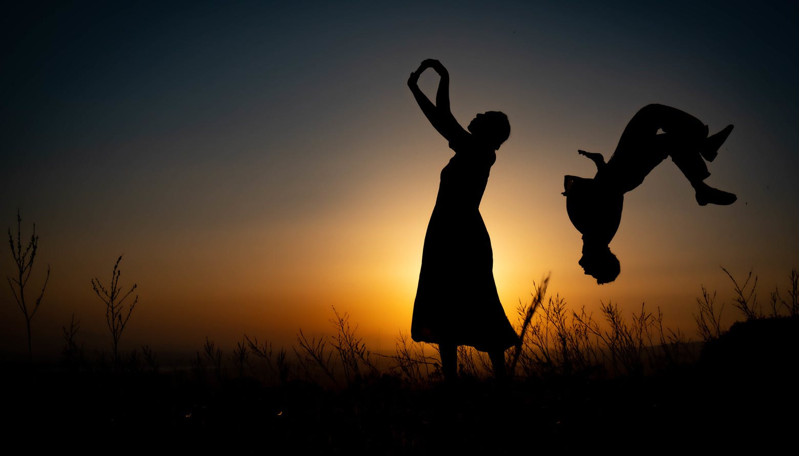At sunset and against the light, a woman stands in the middle with her arms pointing upwards. Behind her a man is upside down doing a somersault.