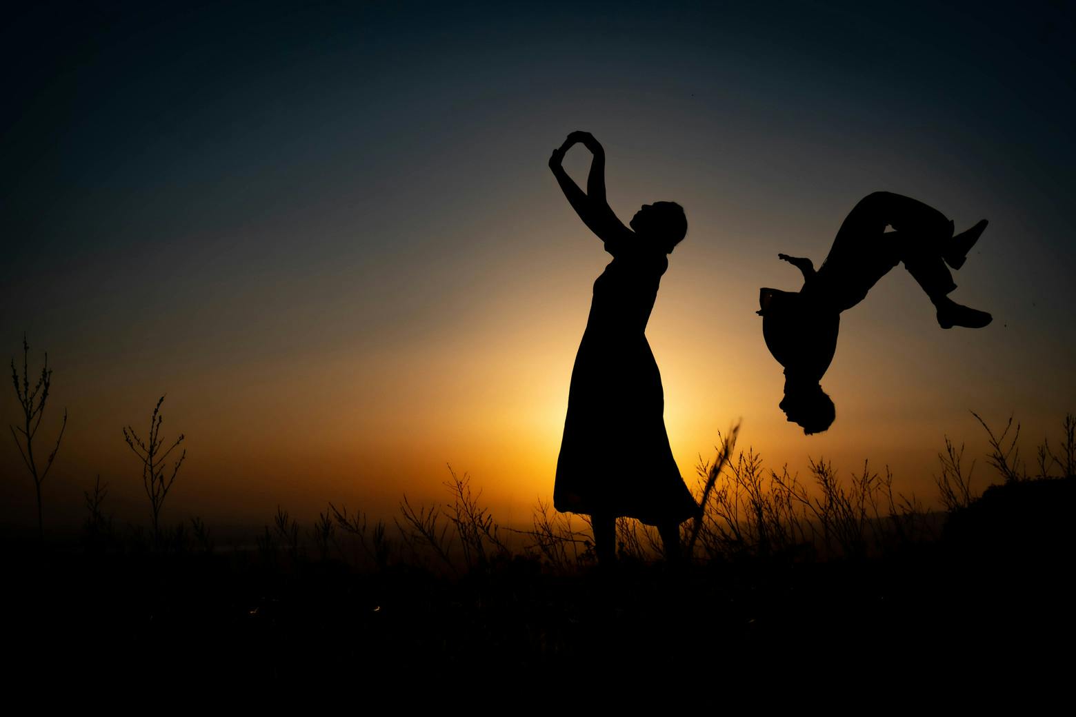 At sunset and against the light, a woman stands in the middle with her arms pointing upwards. Behind her a man is upside down doing a somersault.