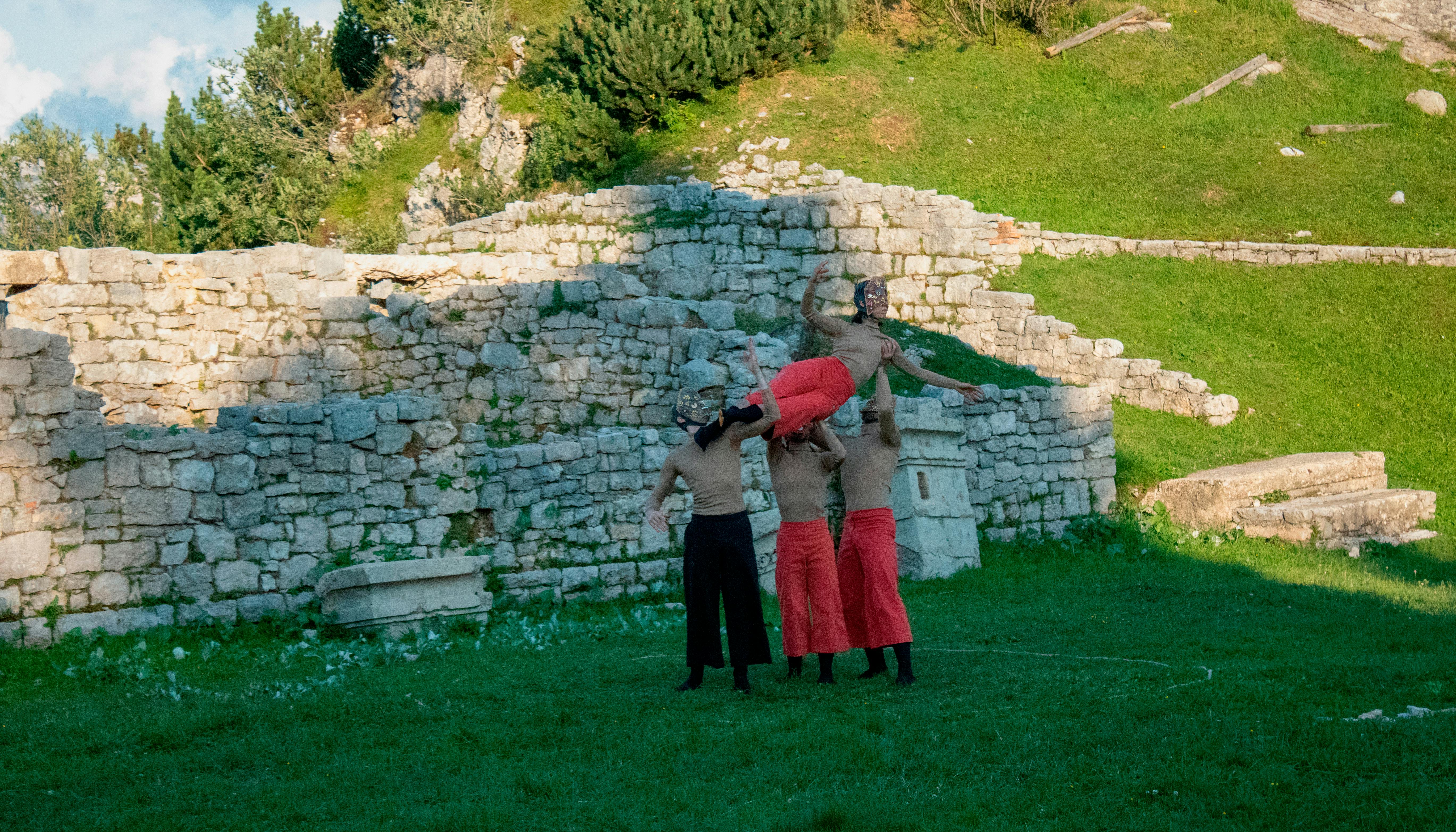 Four performers on a field; one of them is lifted up by the others. They wear masks over their faces. Behind them we see stone ruins and the slope of a hill. It's daytime.
