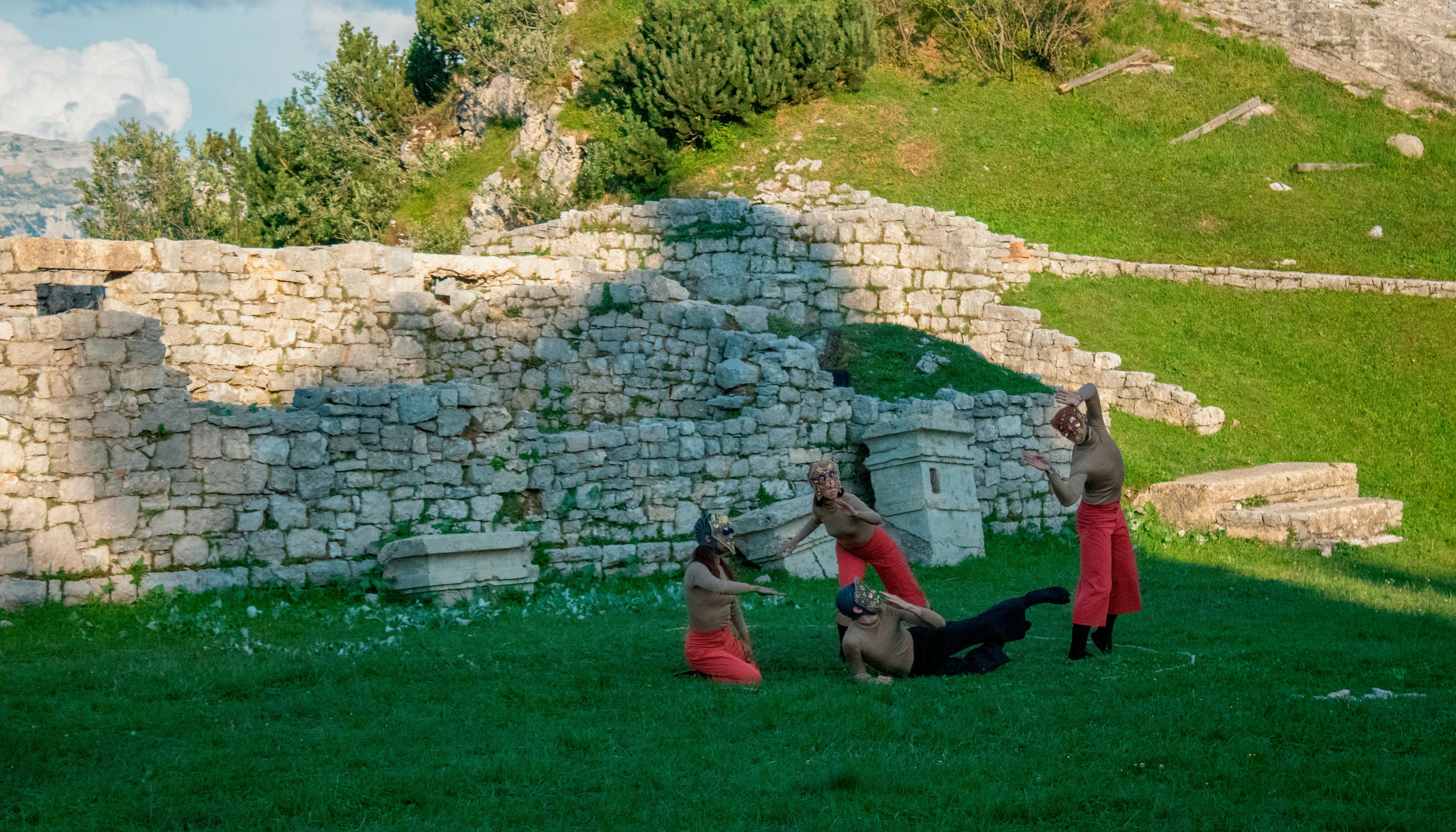 Quattro performer danzano su un prato; indossano pantaloni morbidi, maglie color carne e indossano delle maschere sul viso. Dietro di loro si vedono delle rovine in pietra e il declivio di un'altura. È giorno.