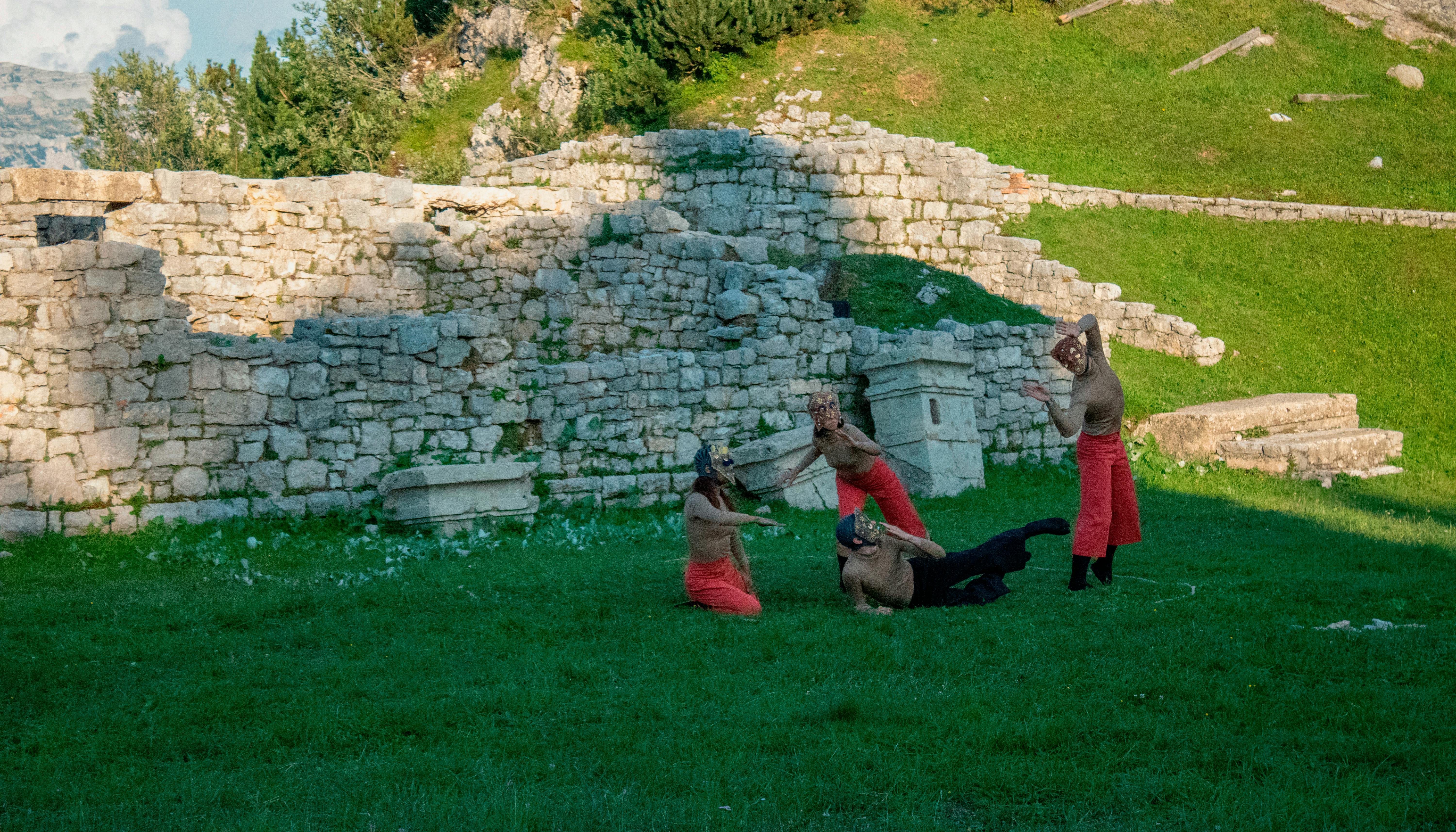 Four performers dance on a lawn; they wear soft pants, flesh-colored shirts, and masks over their faces. Behind them can be seen stone ruins and the slope of a hill. It's daytime.