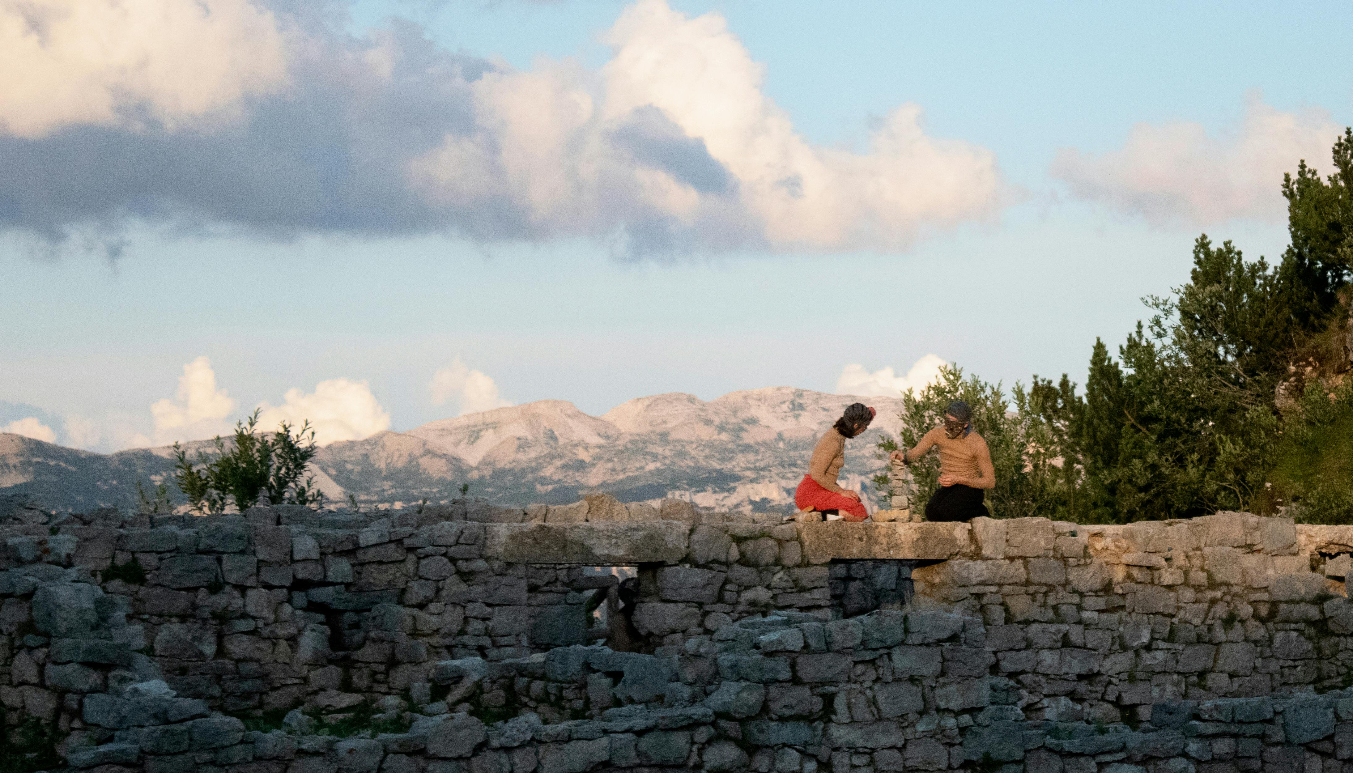 Due performer inginocchiati sopra a un muro in pietra durante lo spettacolo Il mondo altrove. Dietro di loro si apre il panorama: le montagne e il cielo sereno.
