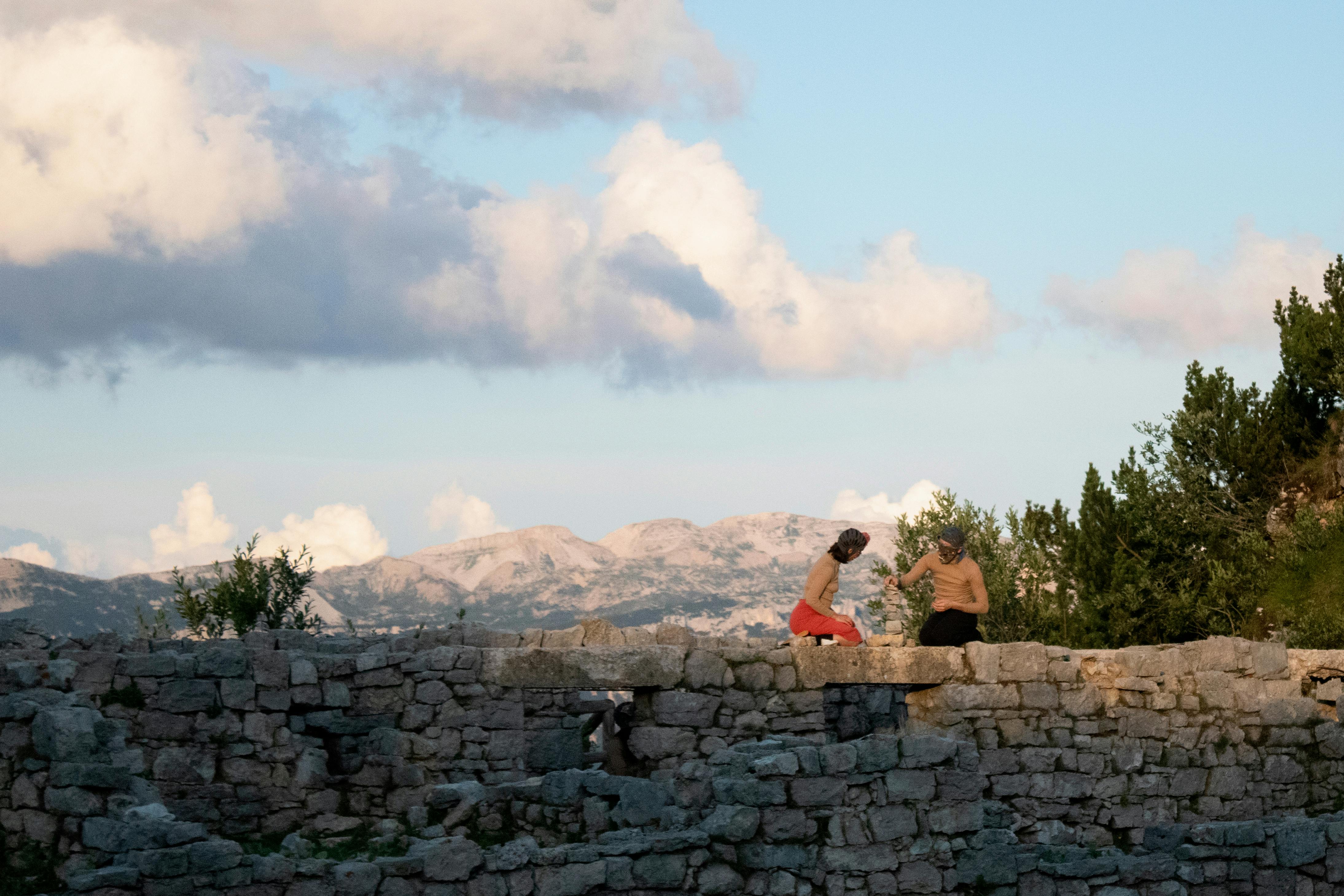 Due performer inginocchiati sopra a un muro in pietra durante lo spettacolo Il mondo altrove. Dietro di loro si apre il panorama: le montagne e il cielo sereno.