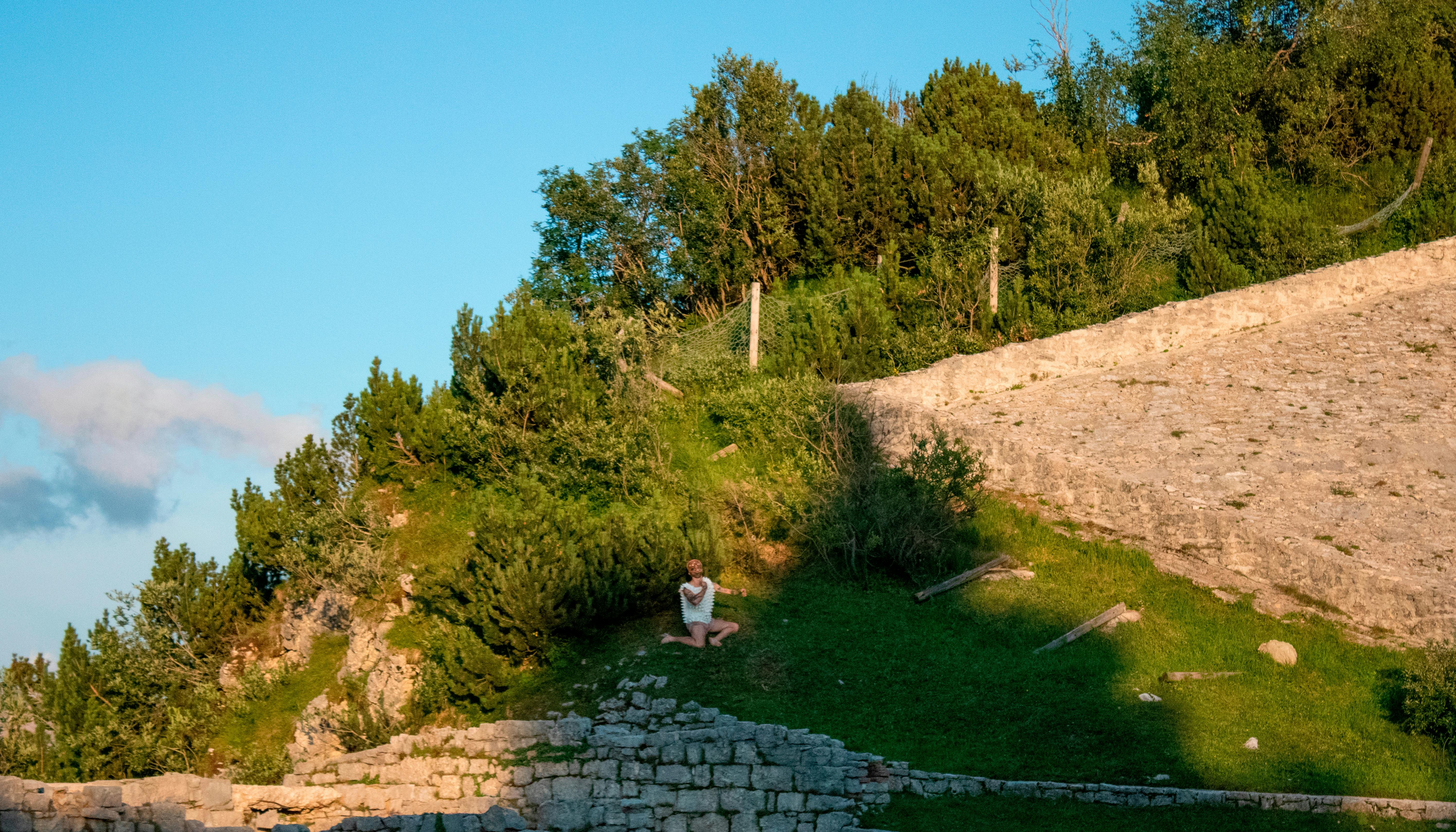 You can see the slope of a hill on a sunny day. Stone ruins are glimpsed. In the landscape, a dancer appears standing out thanks to his white dress.