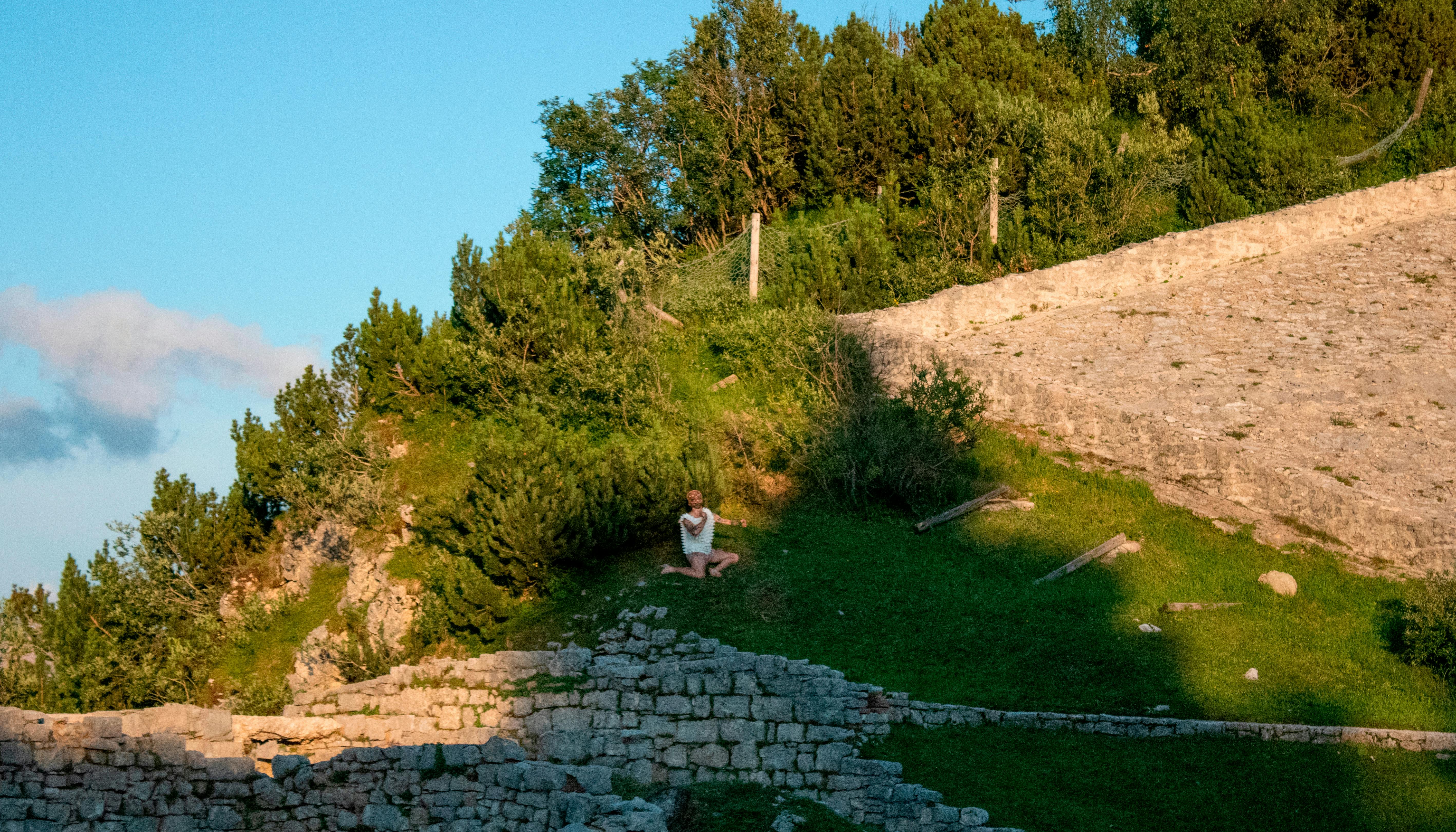 You can see the slope of a hill on a sunny day. Stone ruins are glimpsed. In the landscape, a dancer appears standing out thanks to his white dress.