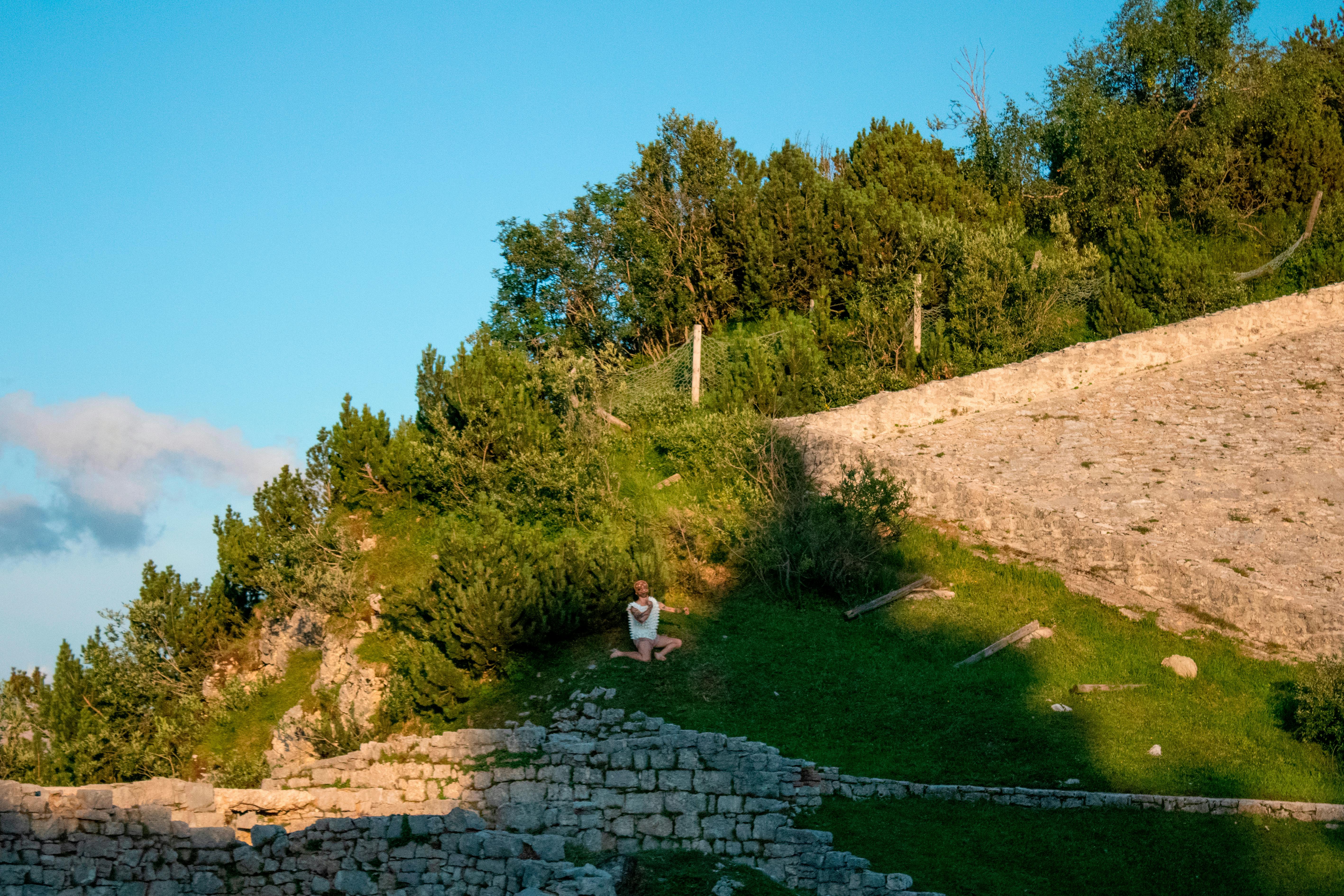 You can see the slope of a hill on a sunny day. Stone ruins are glimpsed. In the landscape, a dancer appears standing out thanks to his white dress.