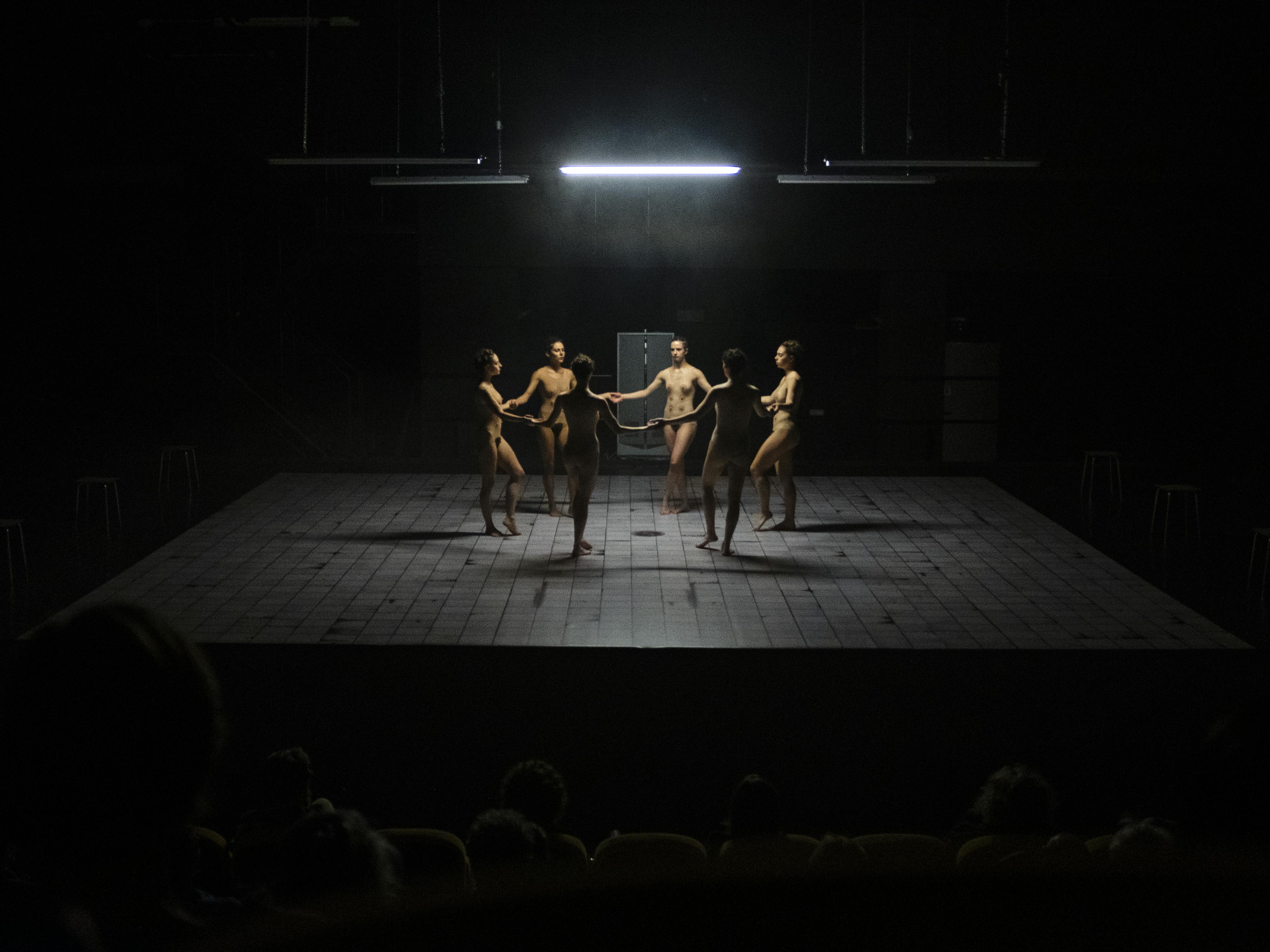 Six female dancers, apparently naked, form a circle on the empty stage. A neon light from above illuminates them.