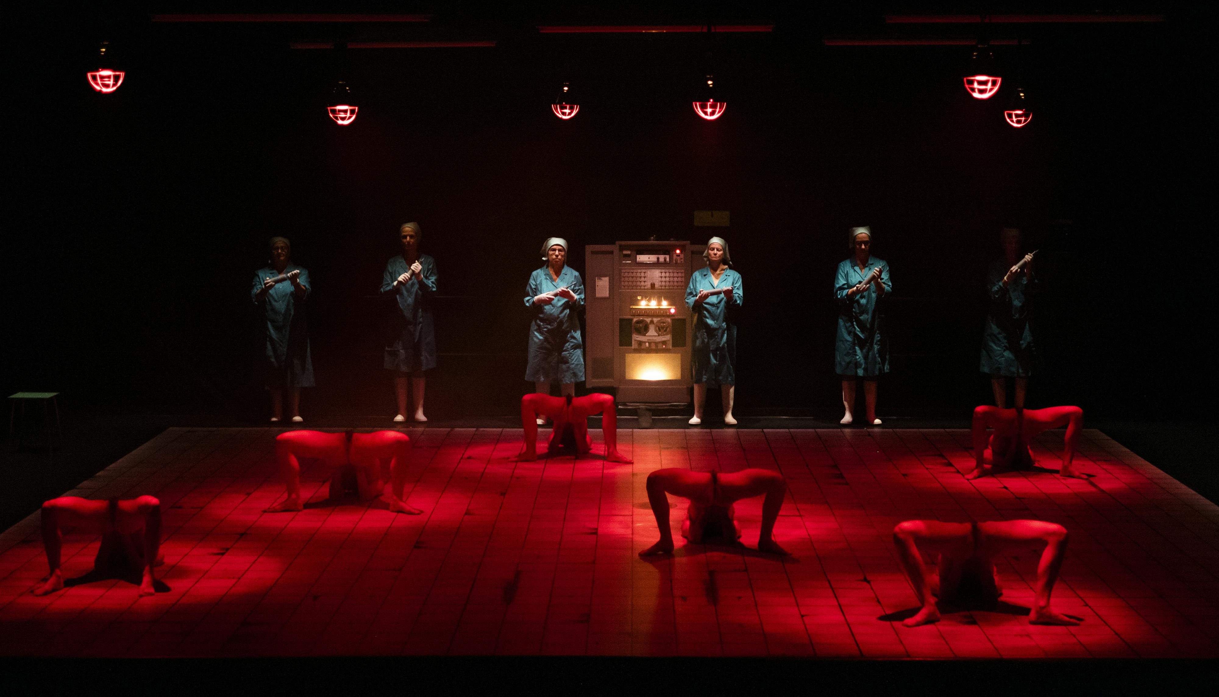 The stage is lit by a dim red light. Six women in lab coats look toward the room from the back of the stage. On the ground in front of them, six decomposed bodies lose their shapes to become something else.