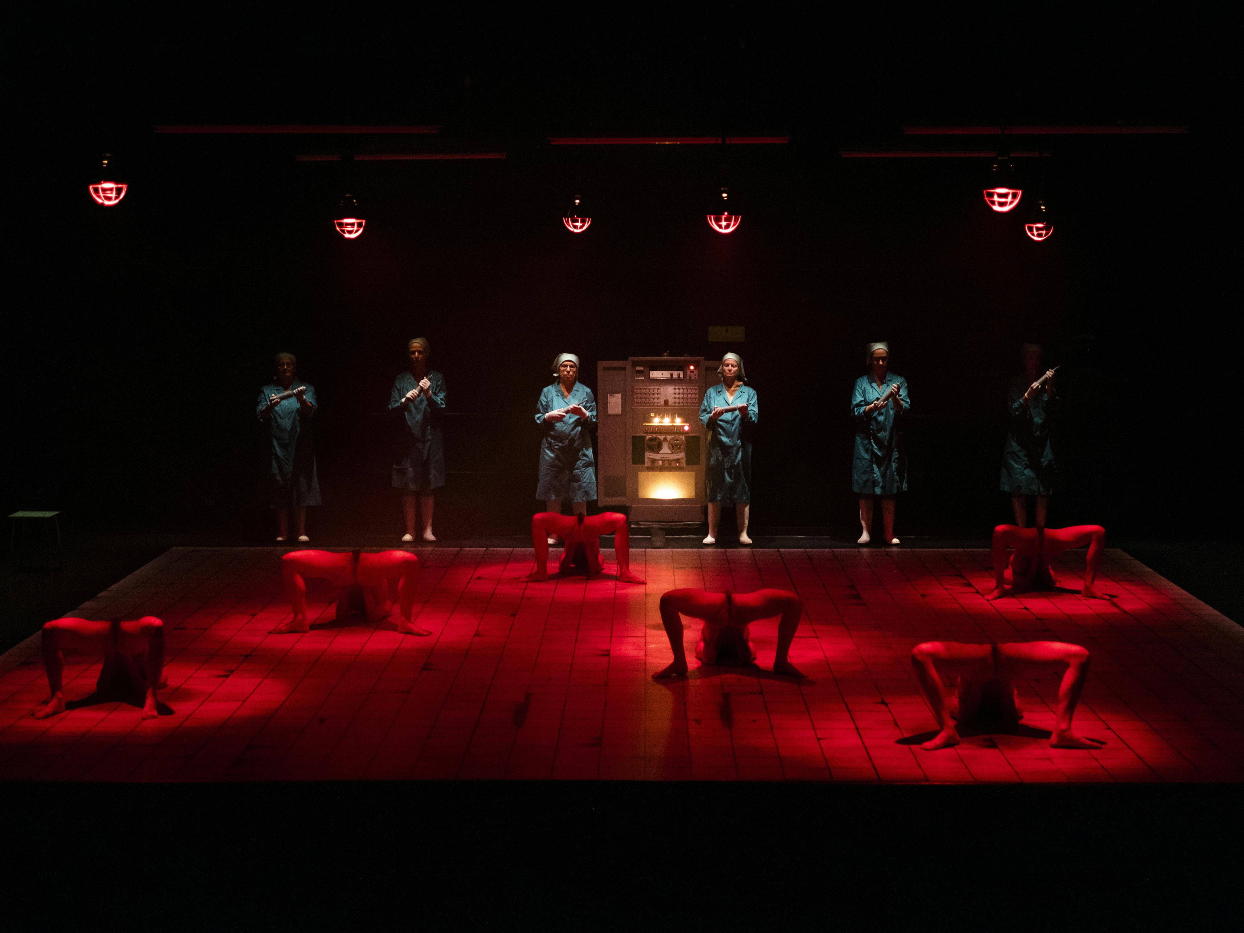 The stage is lit by a dim red light. Six women in lab coats look toward the room from the back of the stage. On the ground in front of them, six decomposed bodies lose their shapes to become something else.