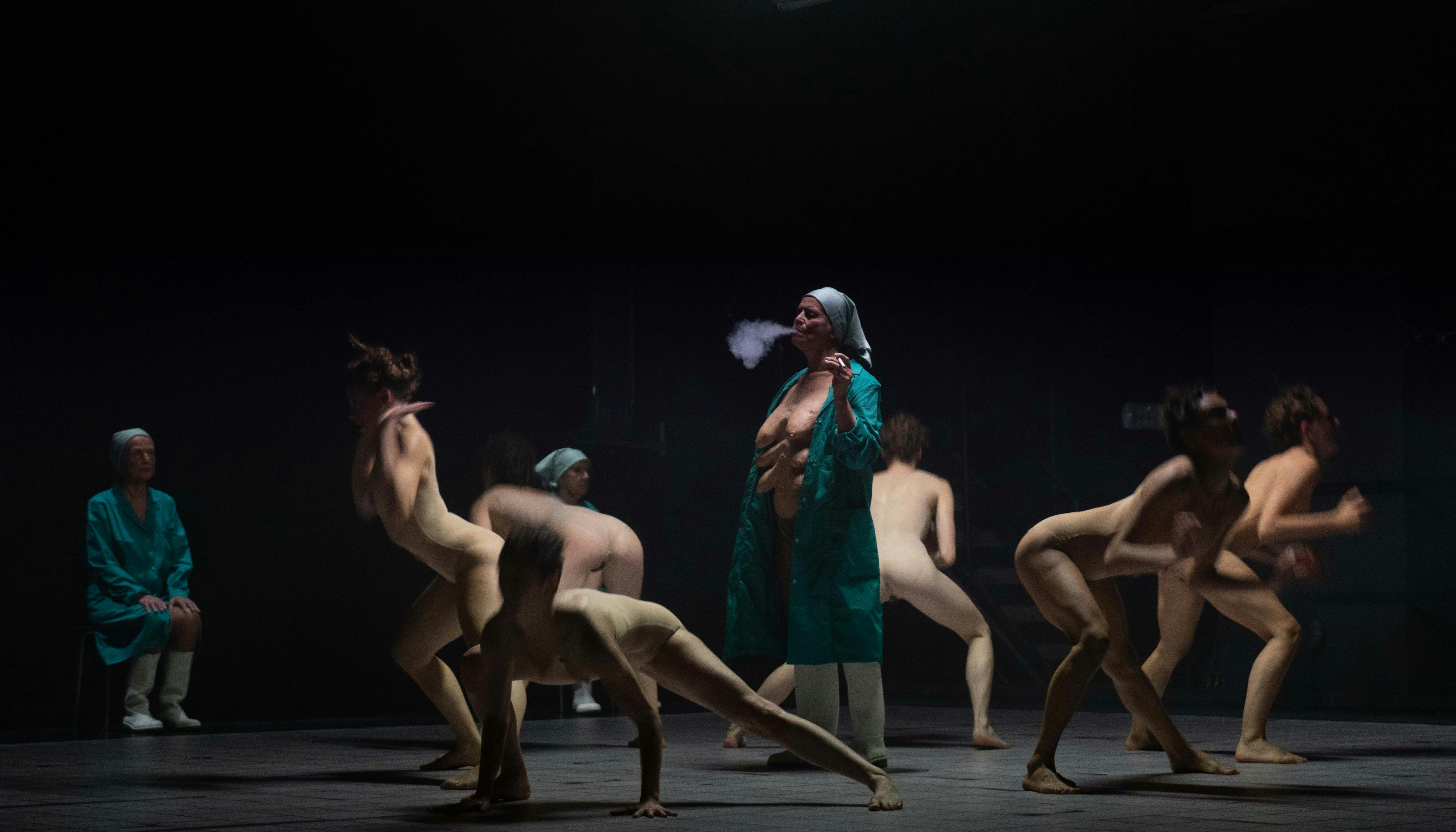 Some naked human bodies dance in a circle, facing outward. In the center is an elderly woman in a lab coat, smoking a cigarette. Under the gown she is naked; multiple pairs of breasts are visible on her chest.