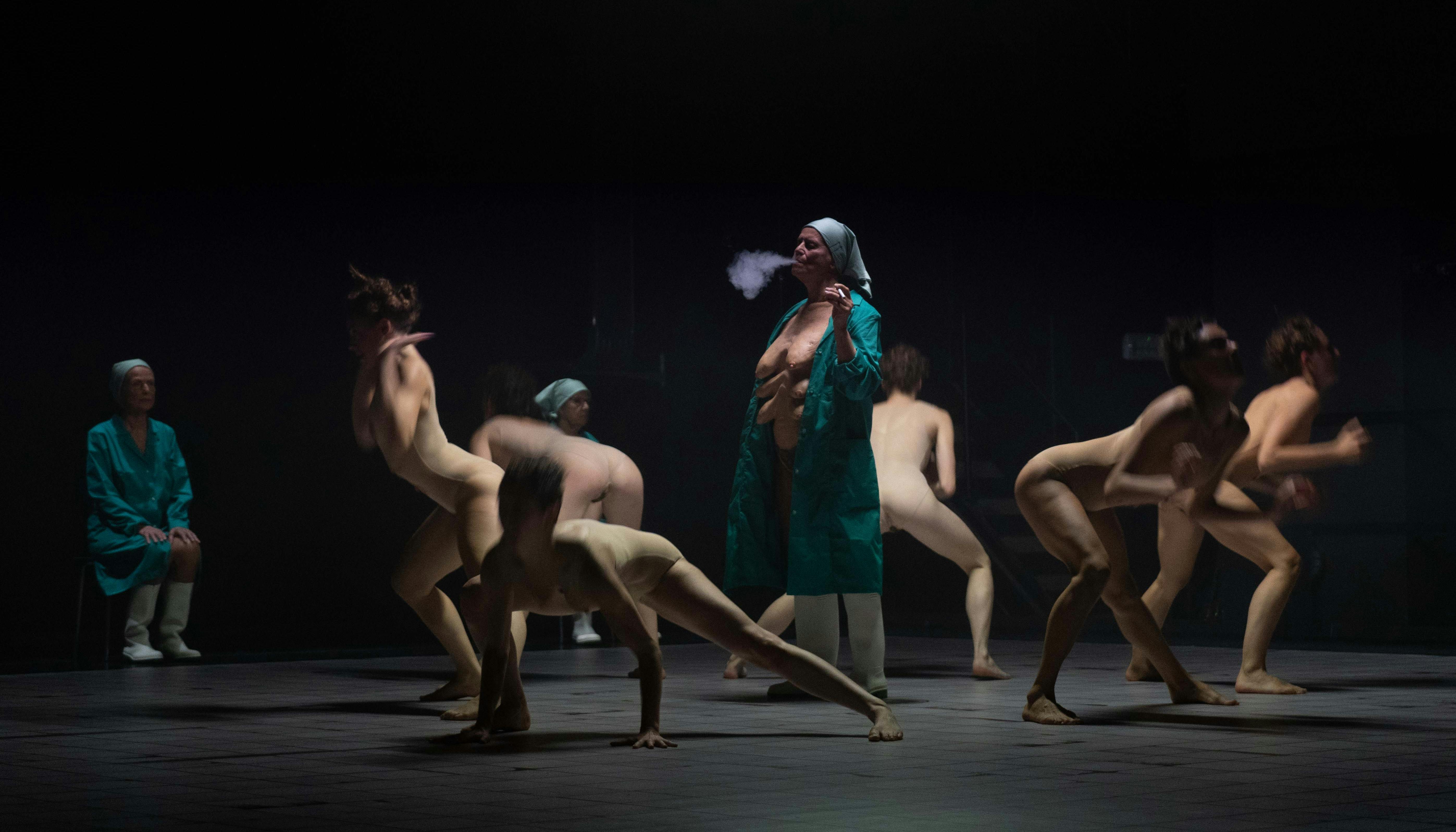 Some naked human bodies dance in a circle, facing outward. In the center is an elderly woman in a lab coat, smoking a cigarette. Under the gown she is naked; multiple pairs of breasts are visible on her chest.