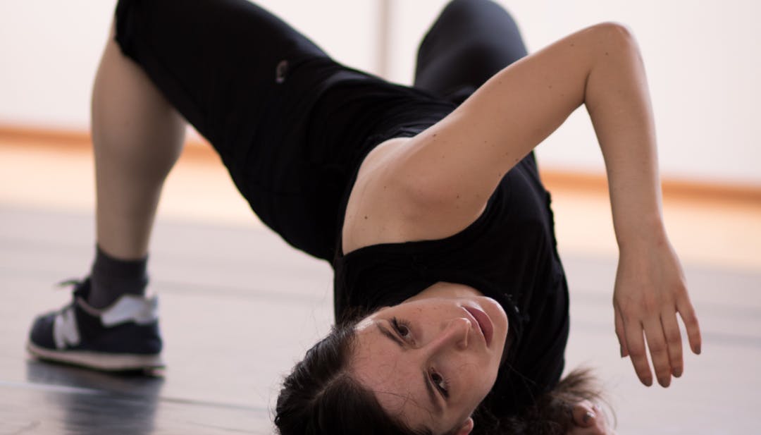 A dancer rehearses in the Studio. She is on the floor, with her feet on the floor, lifting her pelvis upward and rotating her torso, bearing her weight on her right shoulder.
