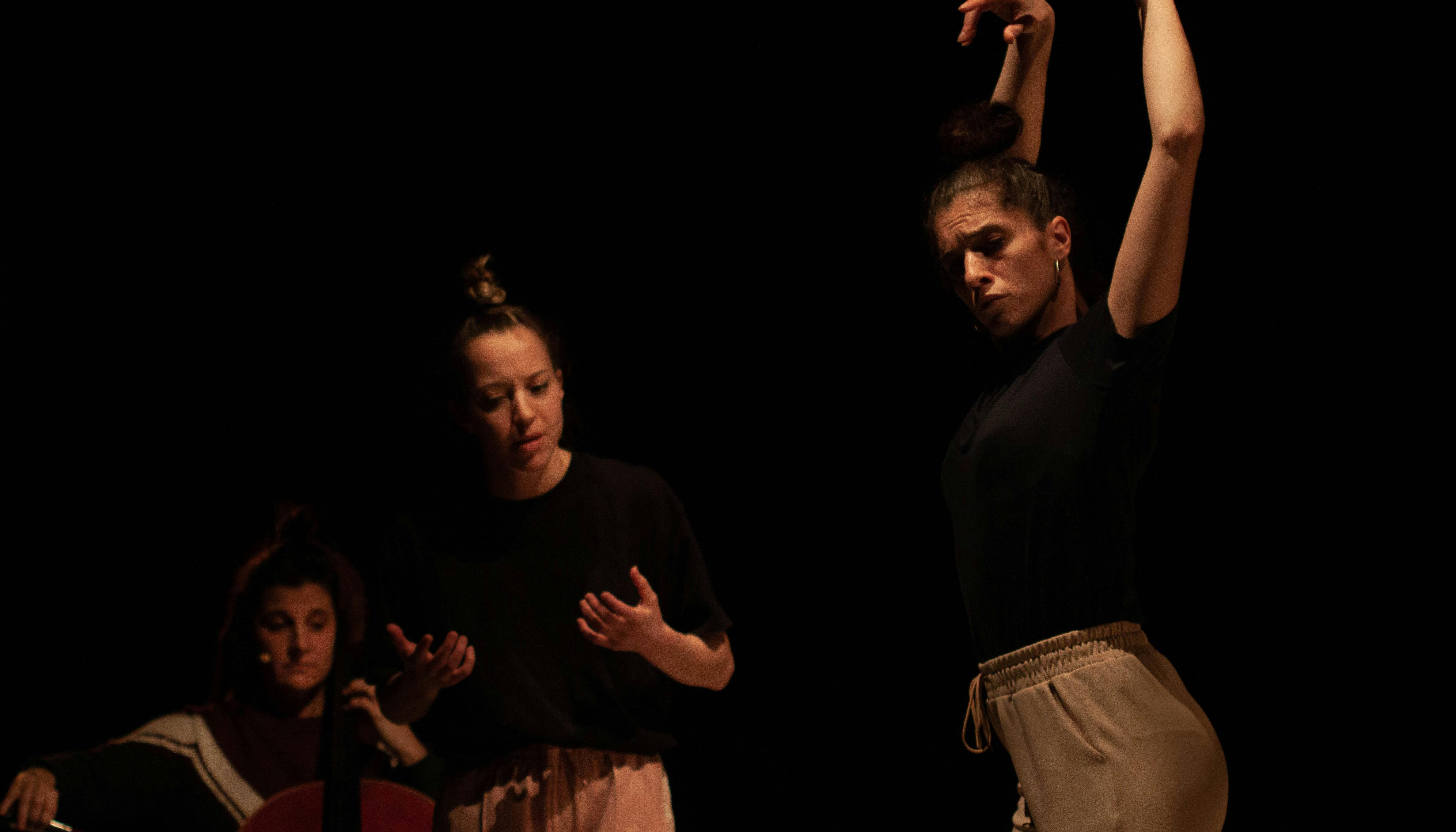 Three performer in rehearsal. In the foreground a dancer with her arms raised upward and her back slightly arched; just behind a second dancer with her hands raised to chest height. In the back a cellist, seated, plays her instrument.