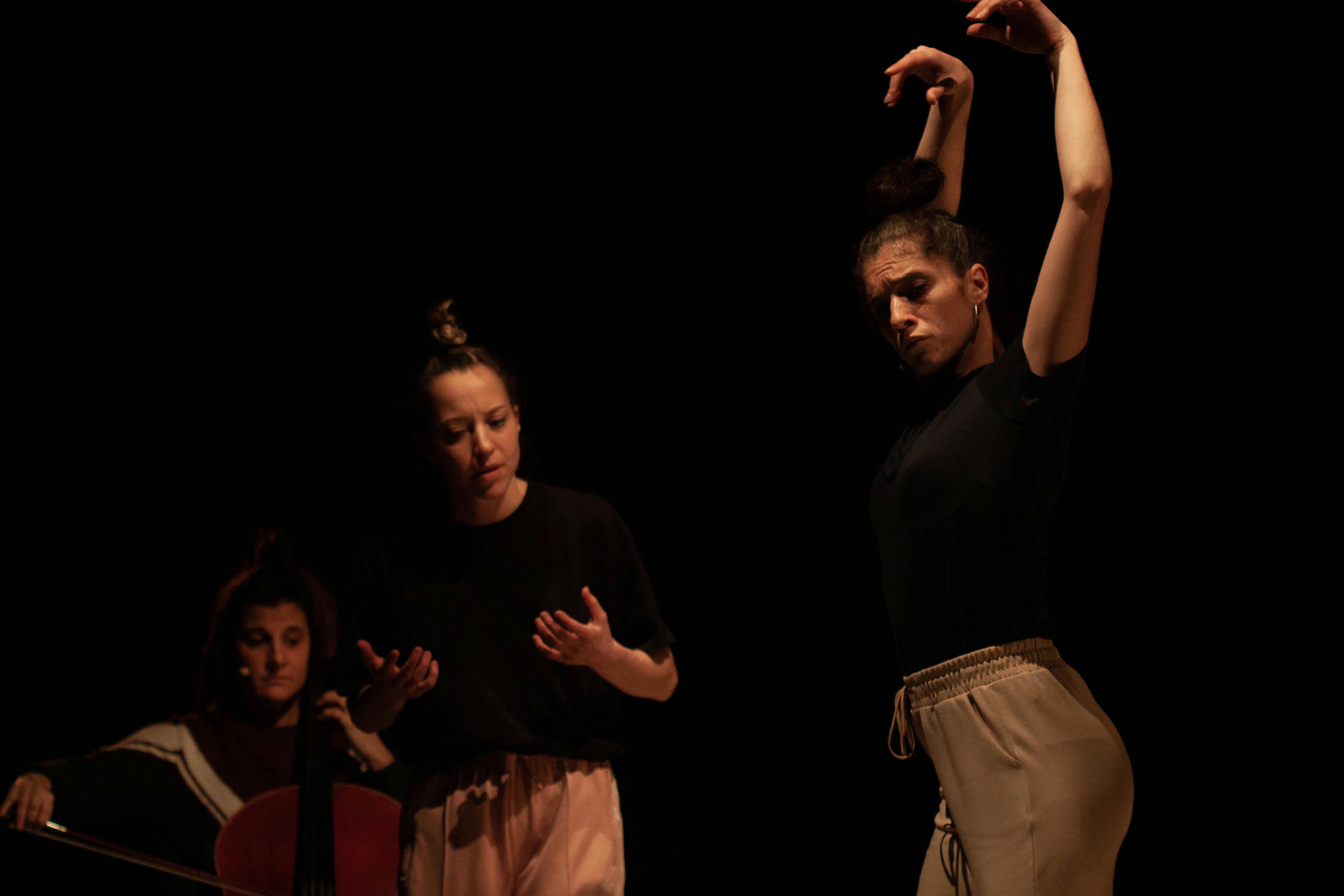 Three performer in rehearsal. In the foreground a dancer with her arms raised upward and her back slightly arched; just behind a second dancer with her hands raised to chest height. In the back a cellist, seated, plays her instrument.